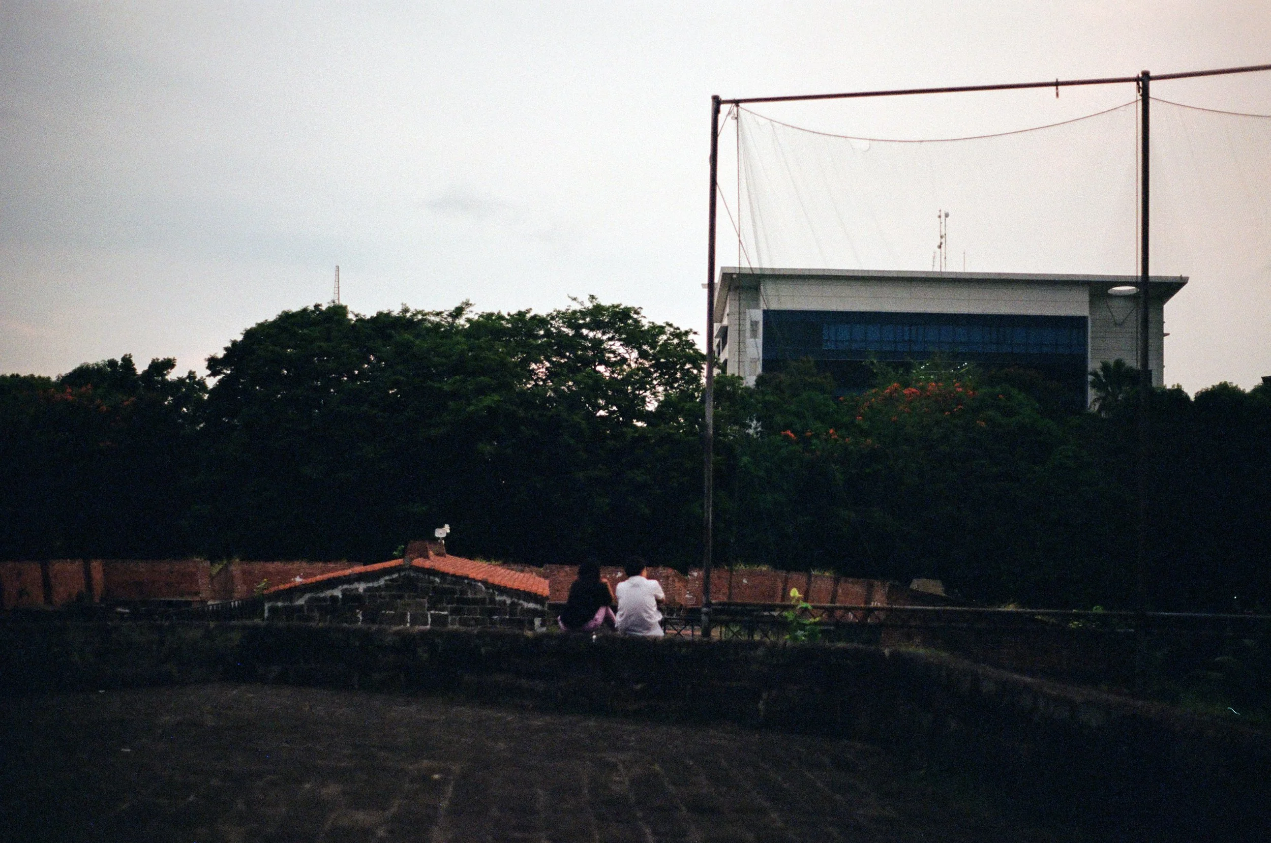 Two people sitting on a rooftop ledge overlooking trees and a large building in the background during dusk.
