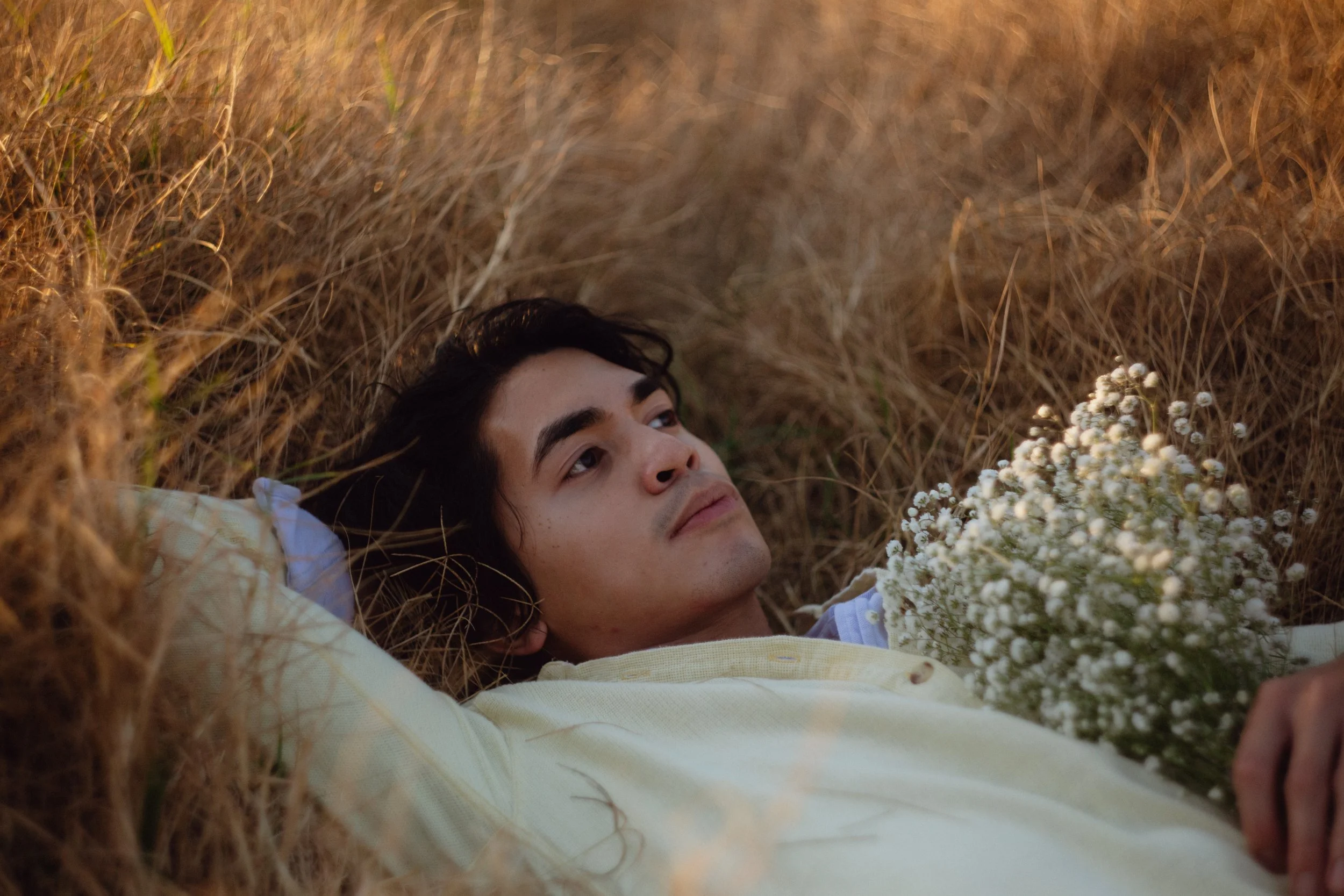 A young man with dark hair and wearing a light yellow shirt lying on his back in a field of tall, dry grass, holding a bouquet of small white flowers.