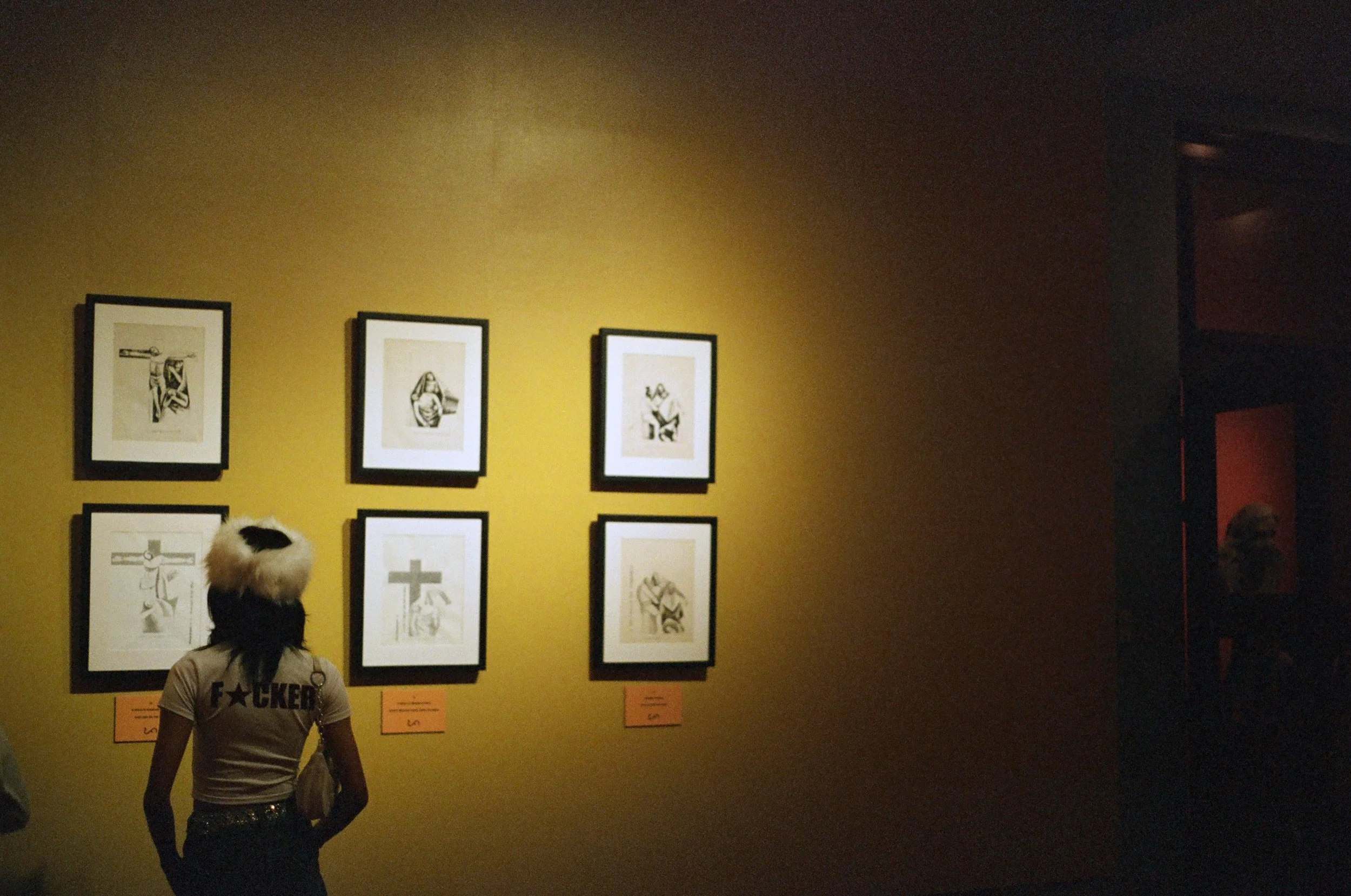 A person with a fur hat and a shirt that says 'F**KER' looks at six framed black-and-white illustrations of the crucifixion on a yellow gallery wall, with numbered orange labels below.