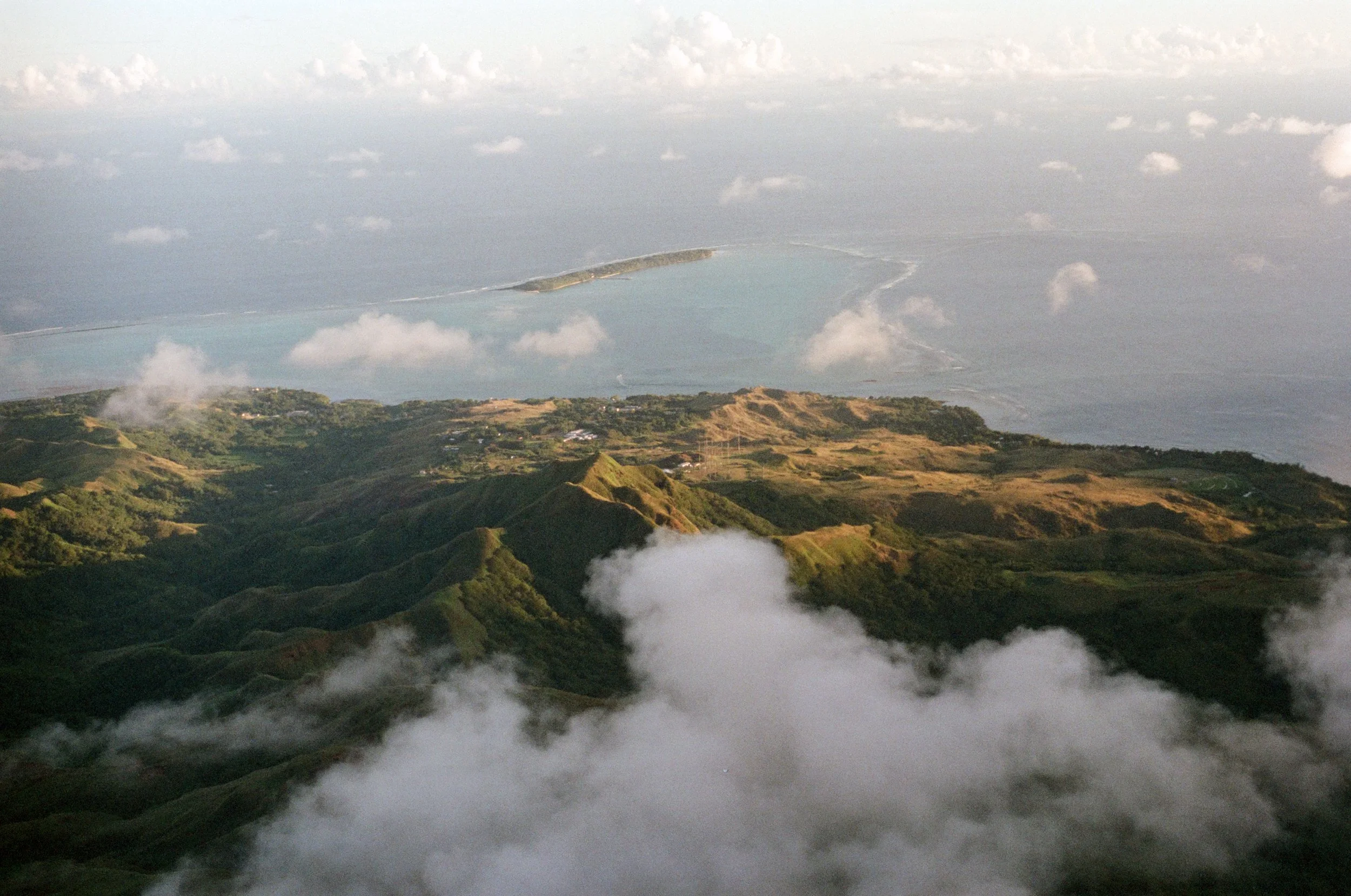 Aerial view of green hilly landscape with clouds and a coastal island in the distance.