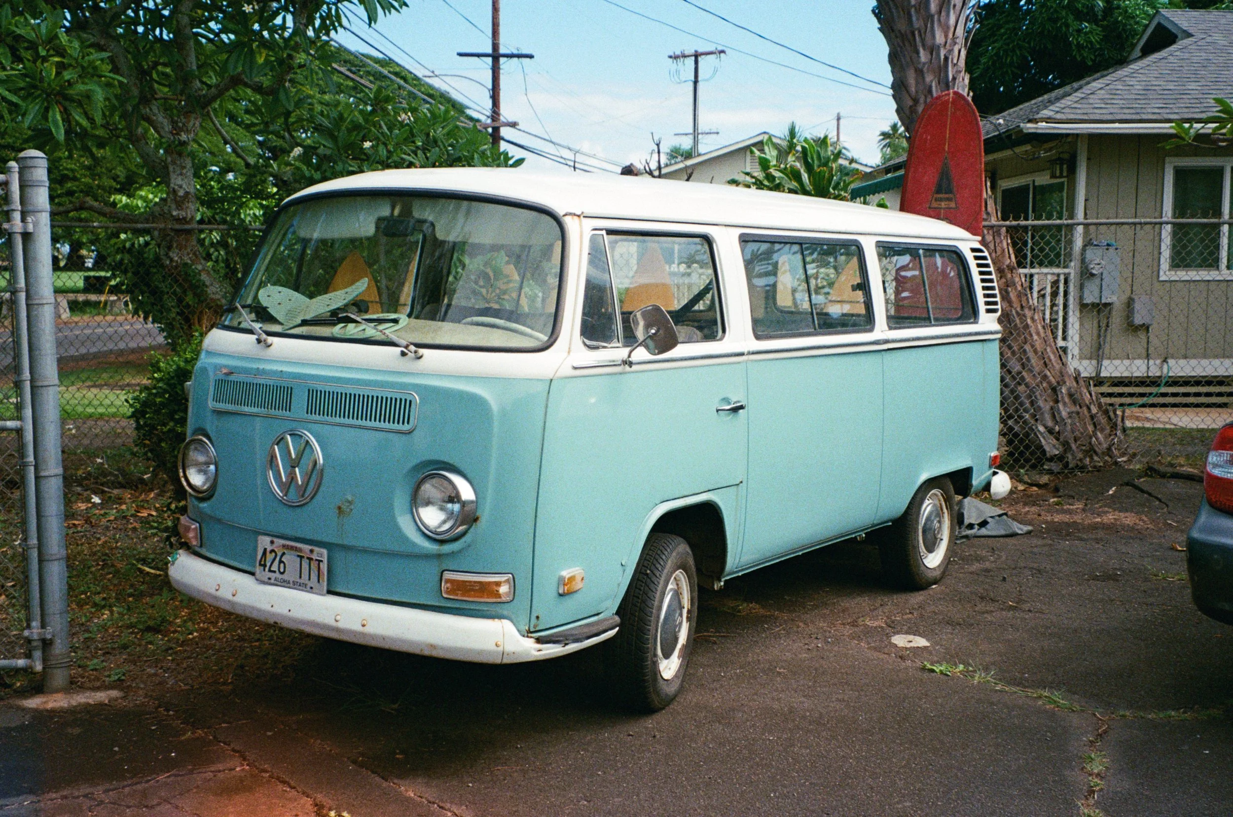 A vintage light blue and white Volkswagen Type 2 van parked on a street with a surfboard behind a tree in the background.