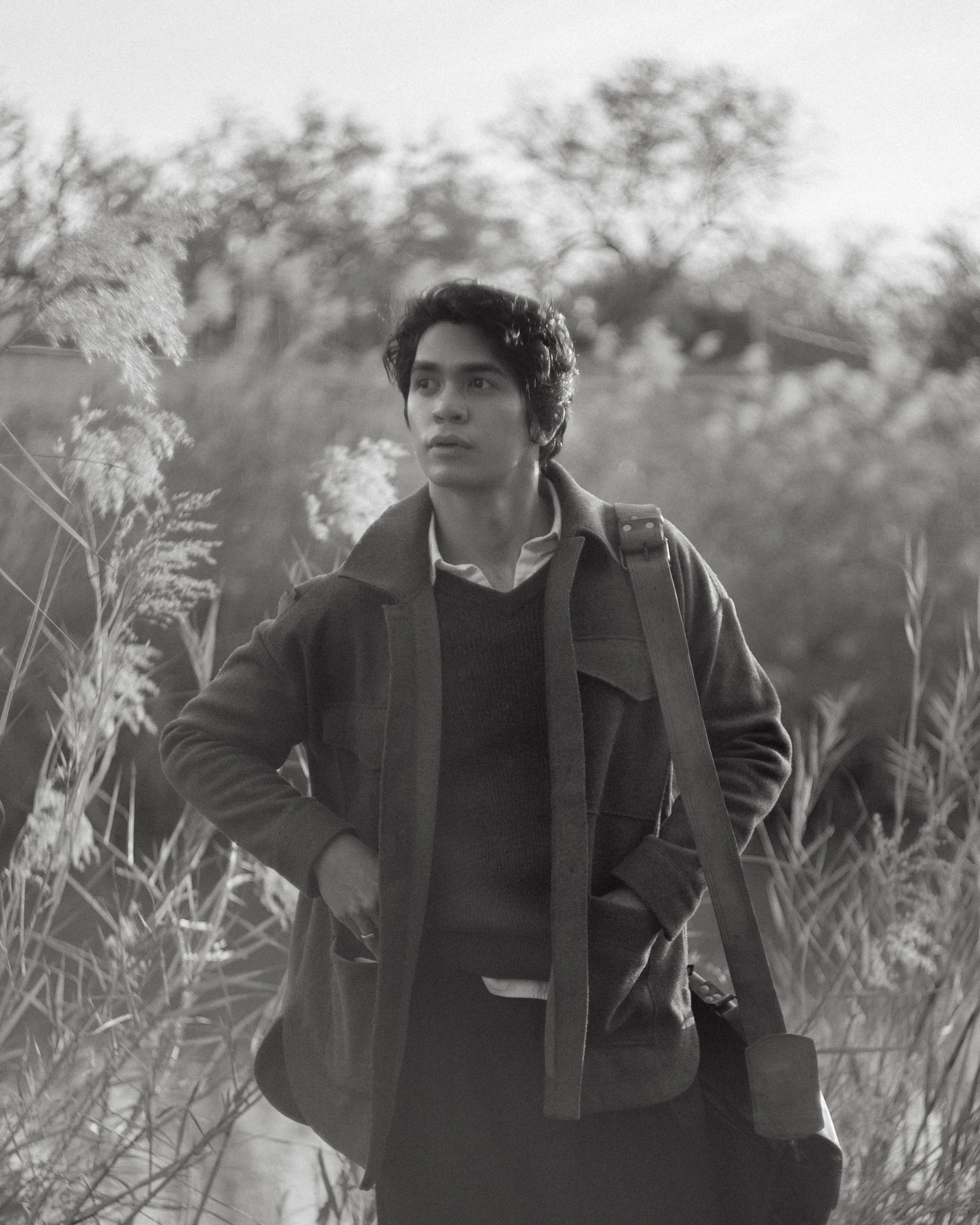 A young man outdoors in a natural setting with tall grass and trees, looking to the side with a contemplative expression, wearing a coat and carrying a shoulder bag.