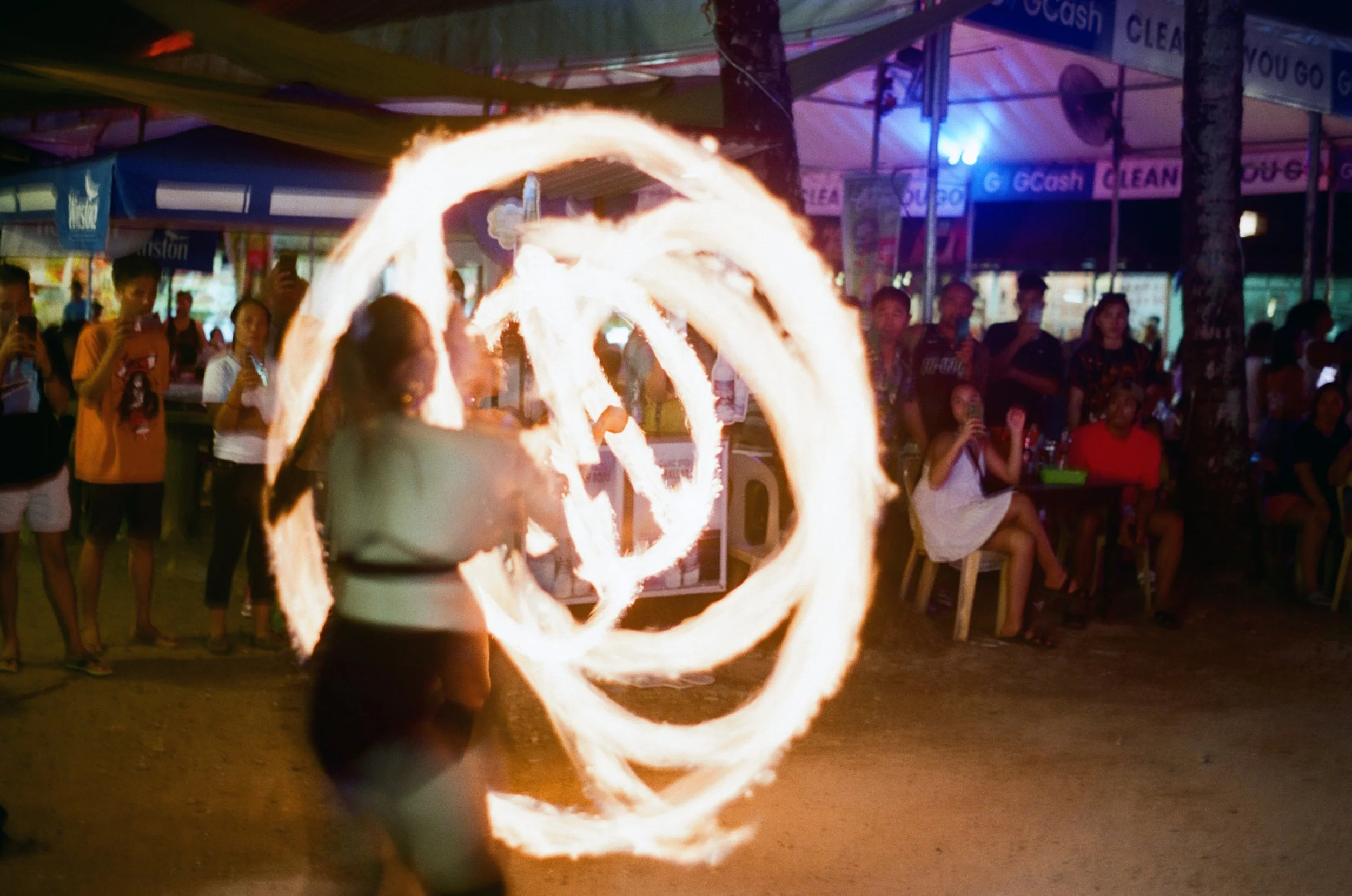 A fire performer creating a fire hoop performance at night in front of a crowd at an outdoor event, with onlookers taking photos and watching.