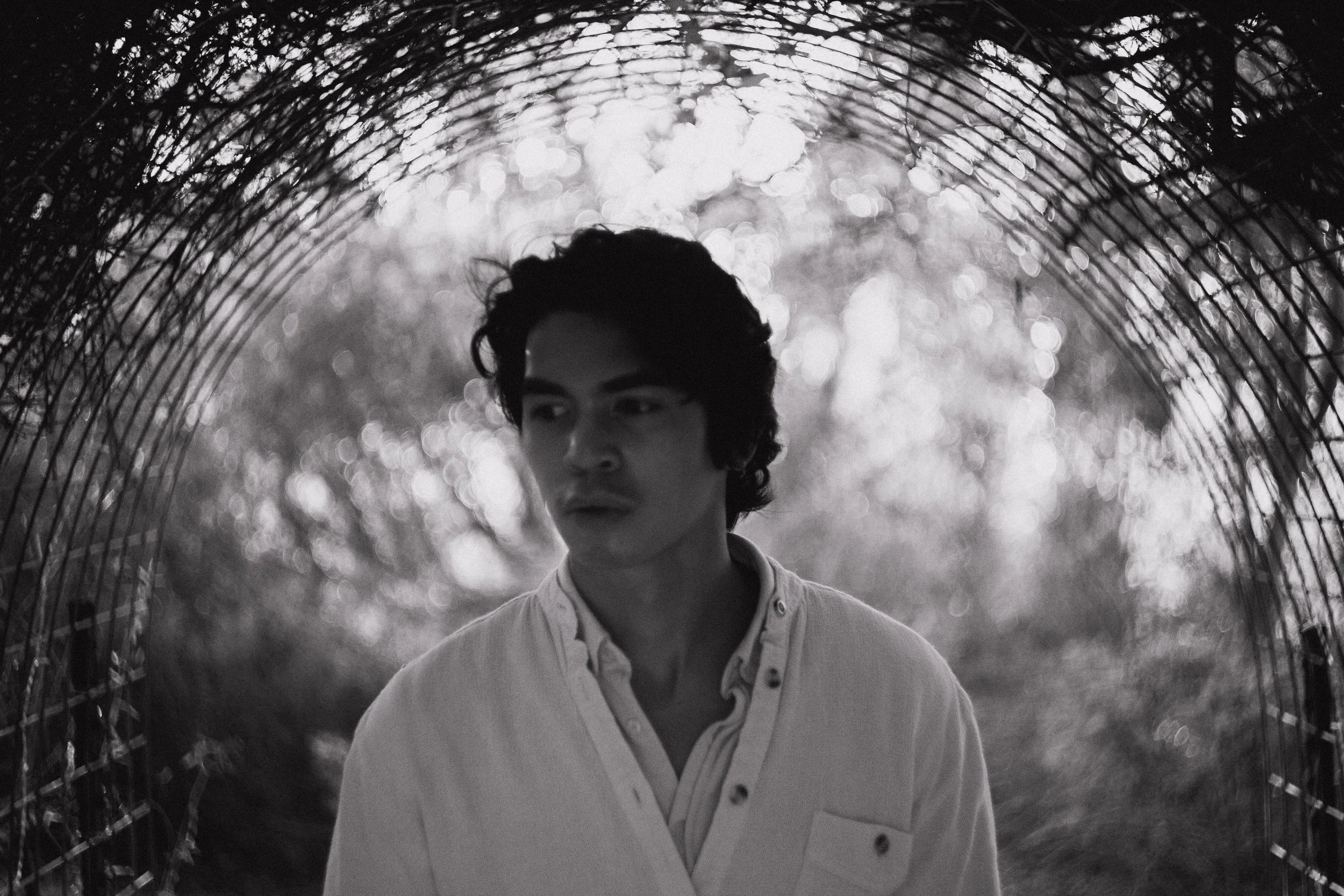 A black and white photo of a young man with dark, wavy hair, standing under a curved, metal fence or tunnel, with blurred trees and sunlight in the background.