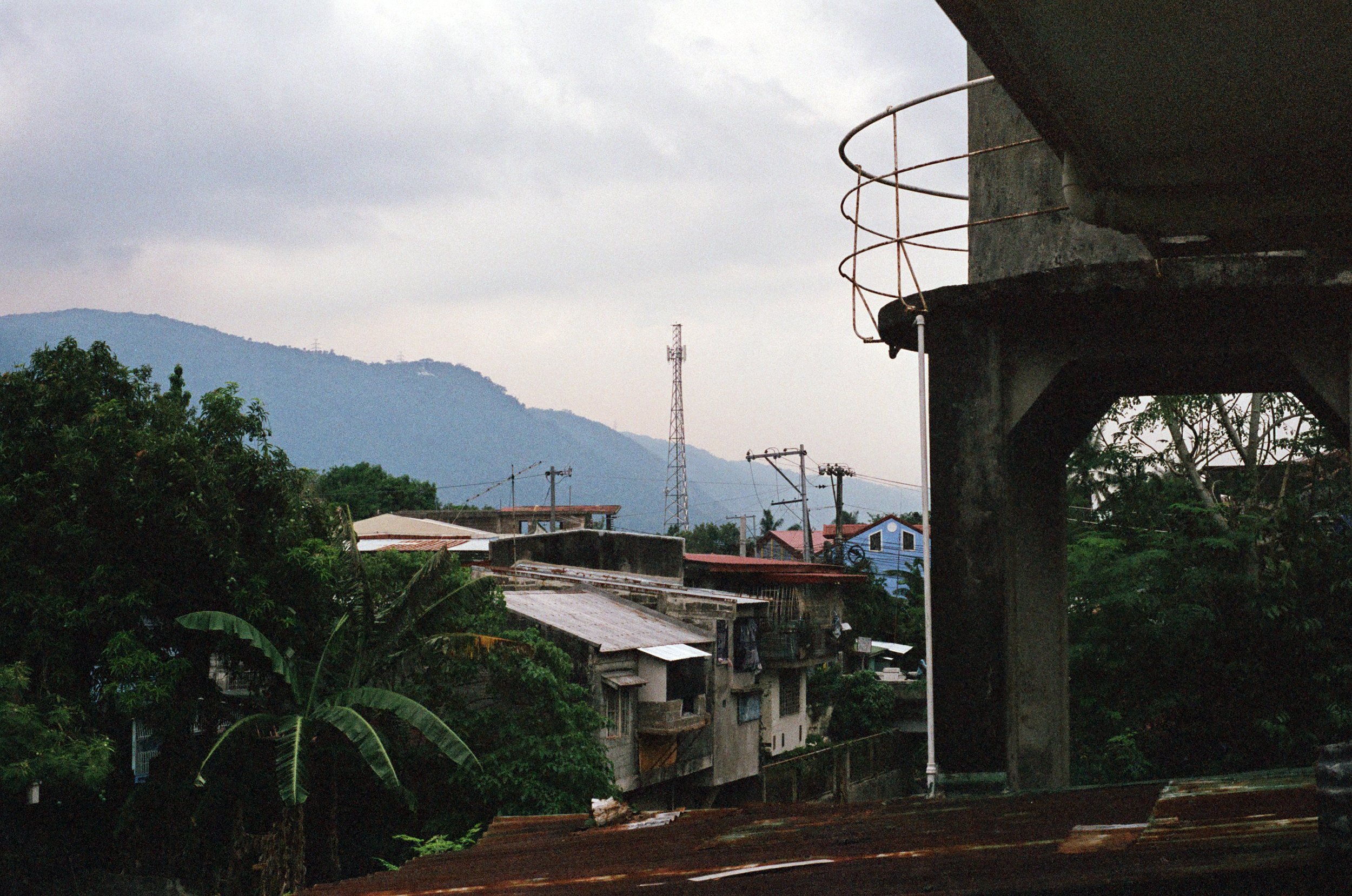 View of a small village with houses, trees, utility poles, and a mountain in the background on a cloudy day.