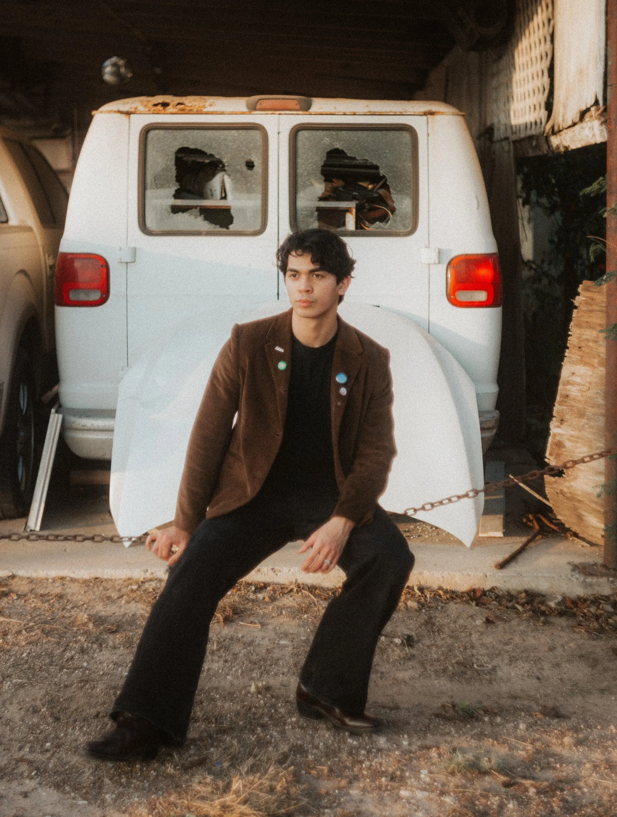 A young man with dark curly hair, wearing a brown blazer and black pants, sitting on a chain in front of a white van with broken windows, outdoors.