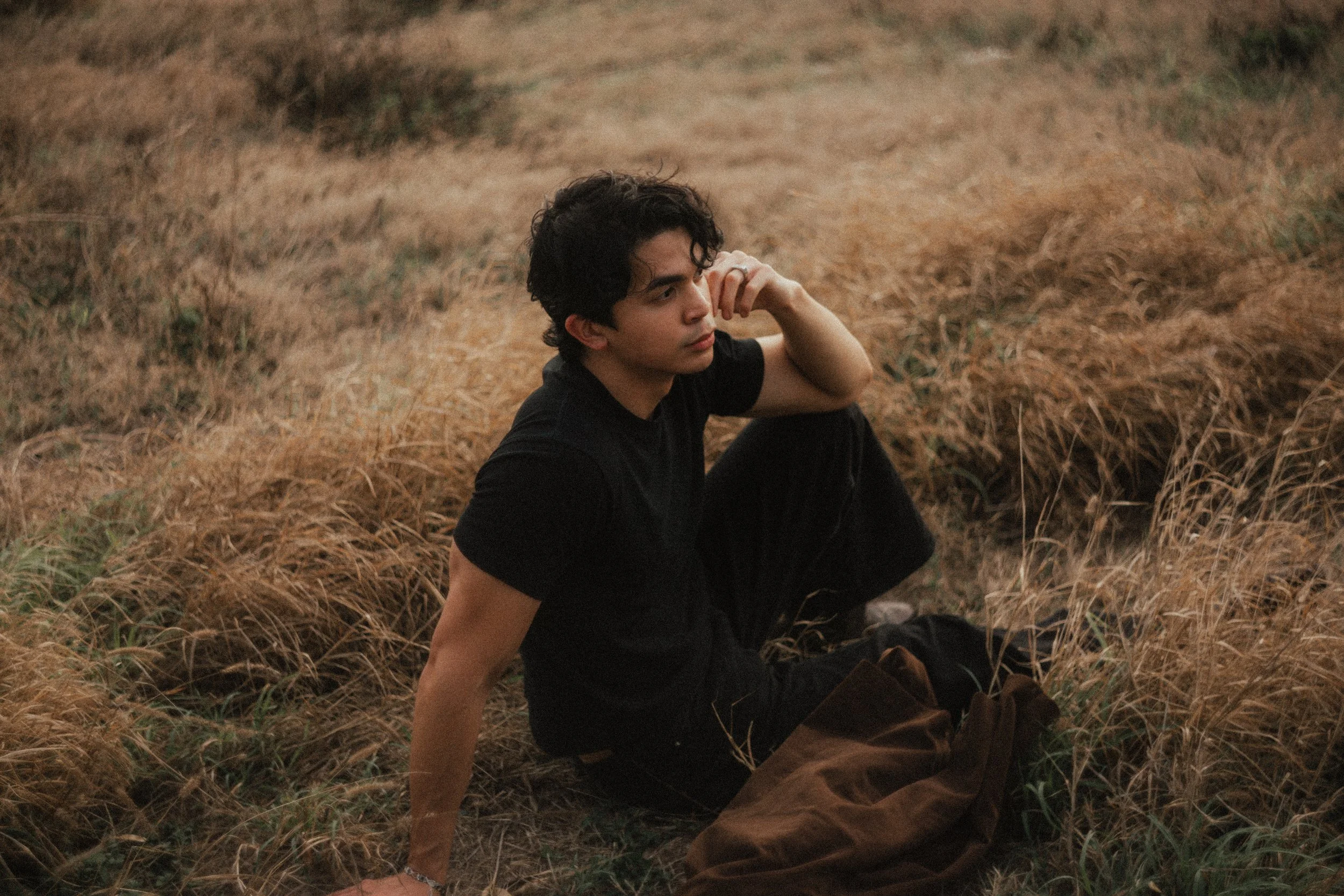 Young man with curly hair sitting on the ground in a field of tall, golden grass, wearing a black t-shirt and dark pants, resting his arm on his knee and looking thoughtful.