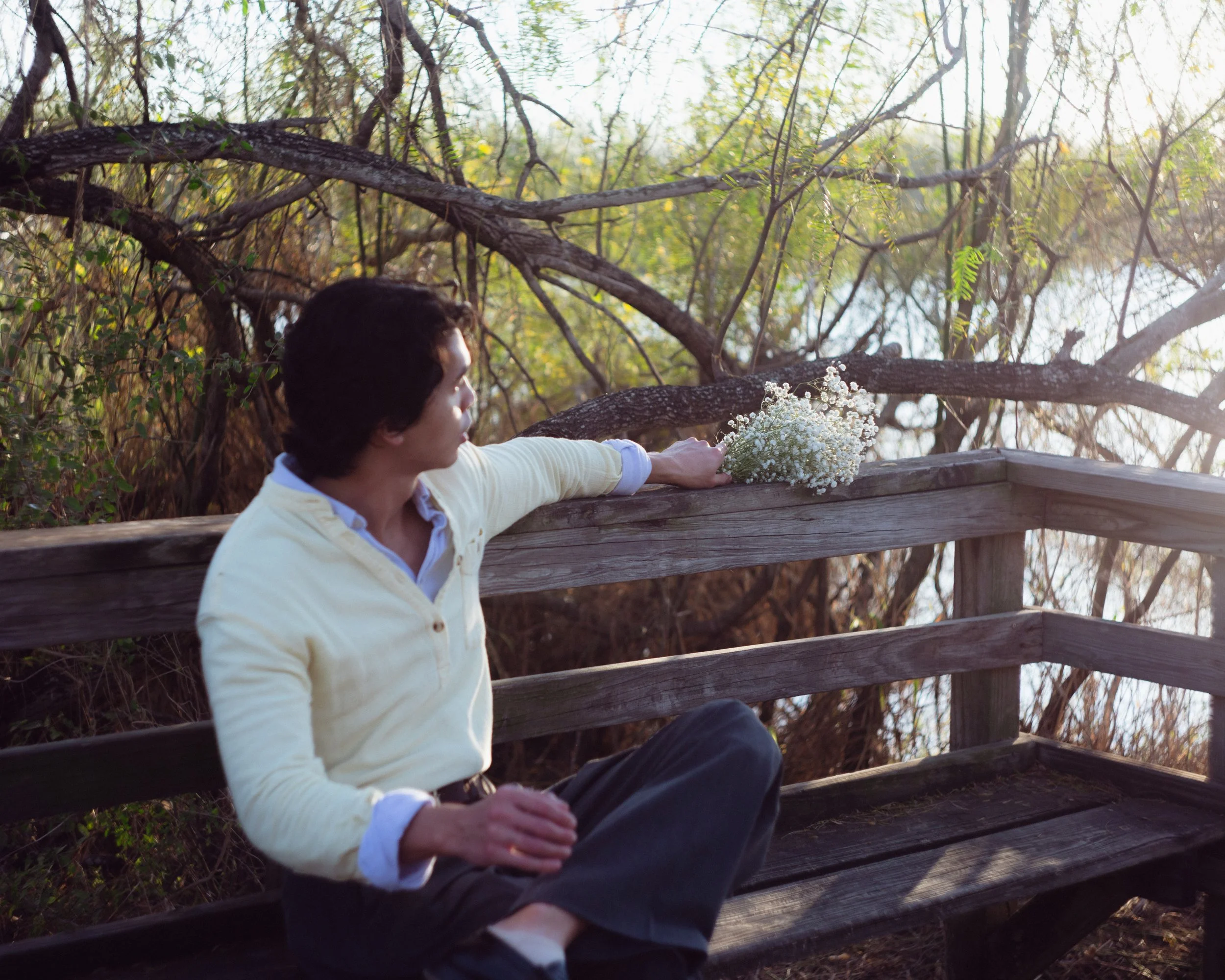 A man sitting on a wooden bench outdoors, reaching with his arm extended to a bouquet of white flowers on the bench, overlooking a water body with trees in the background.