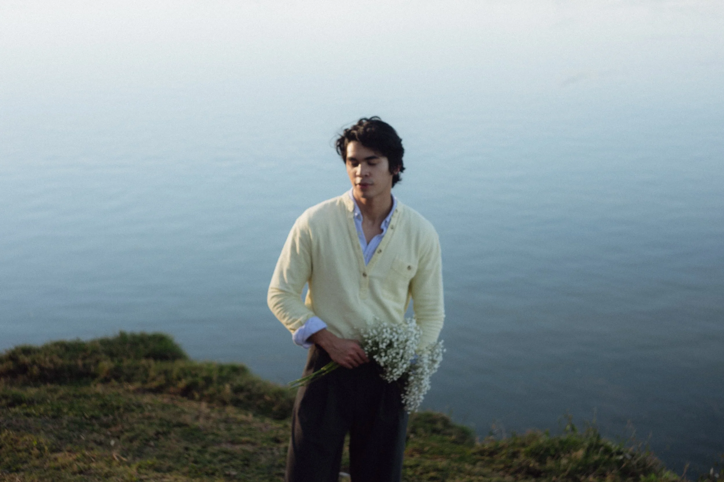 A young man with dark hair standing on a grassy area near a body of water, holding a bouquet of small white flowers, with a serene water backdrop.