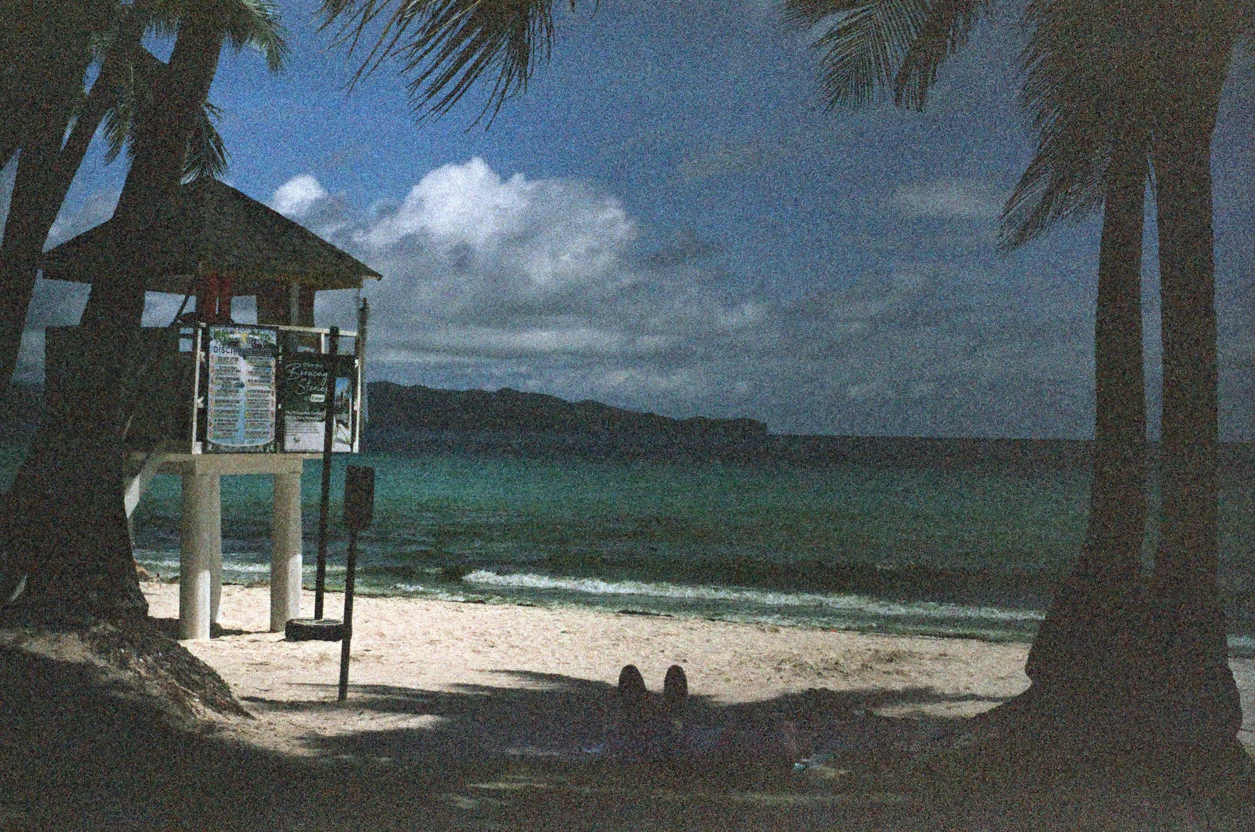 Nighttime view of a beach with two palm trees framing the scene, a person lying on the sand, a lifeguard stand with informational signs, and mountains in the background under a cloudy sky.