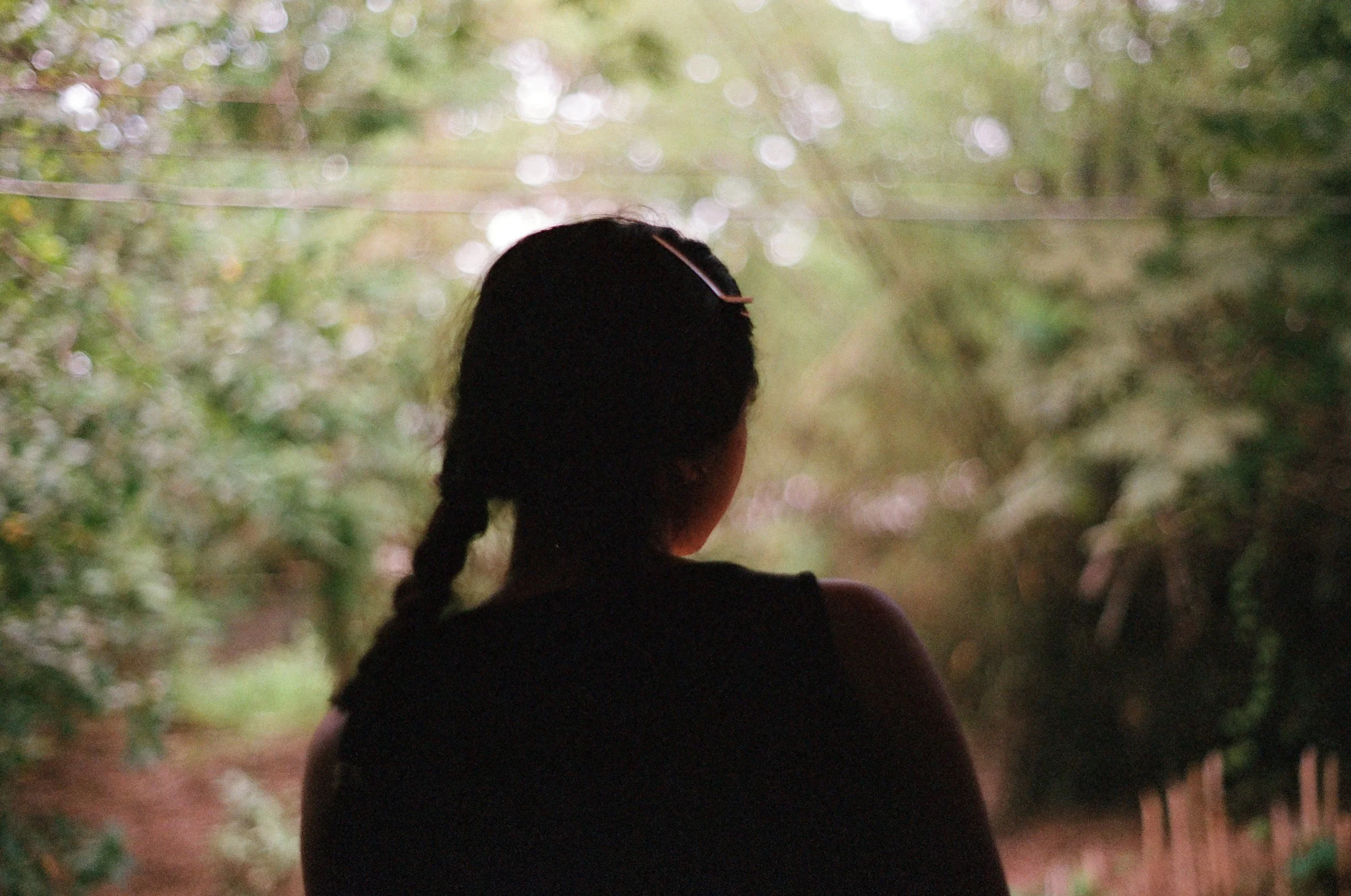 A person with long, dark hair styled in a braid, facing away from the camera, standing in a lush, green outdoor setting with trees and foliage.