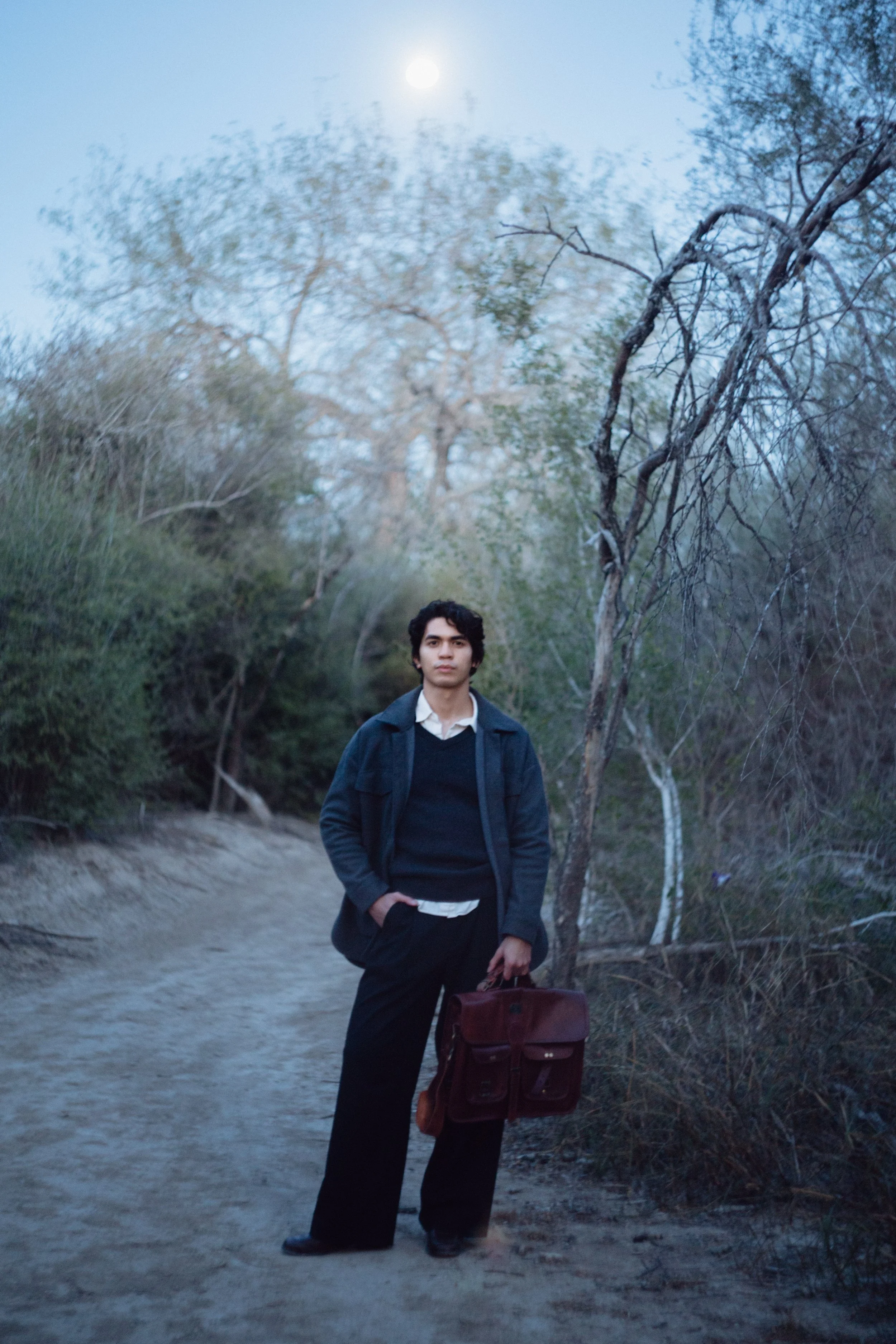 A young man with dark, curly hair stands on a dirt path outdoors, holding a burgundy briefcase in his right hand and wearing a dark blue jacket over a black sweater, with a white shirt underneath. The background features leafless trees and bushes, wi