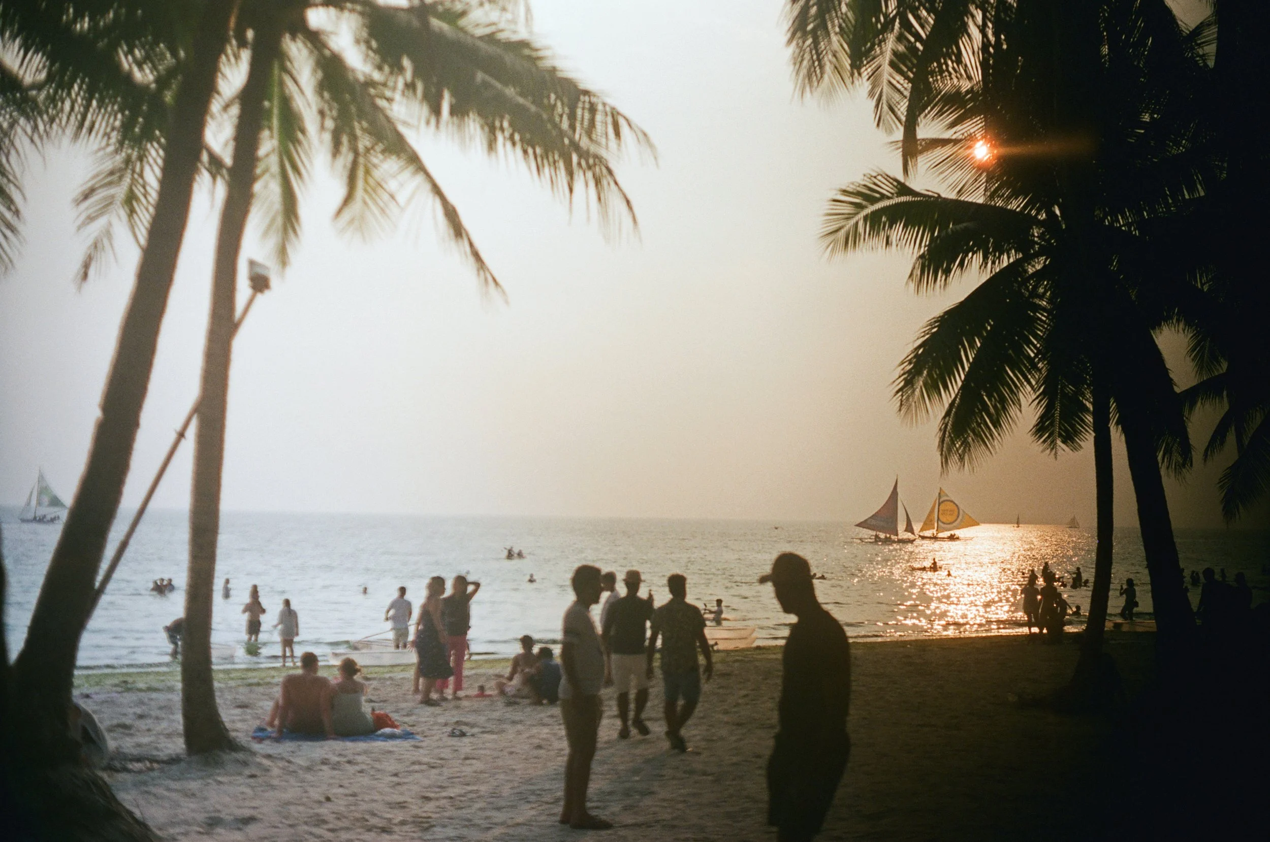 Beach scene at sunset with silhouettes of people walking and sitting under palm trees, sailboats on the water, and the sun reflecting on the ocean.