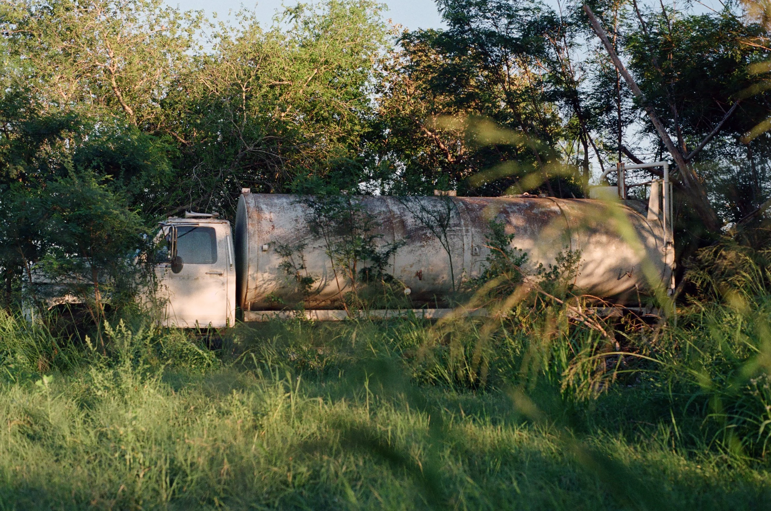 An abandoned, rusty white tanker truck overgrown with tall grass and surrounded by trees.
