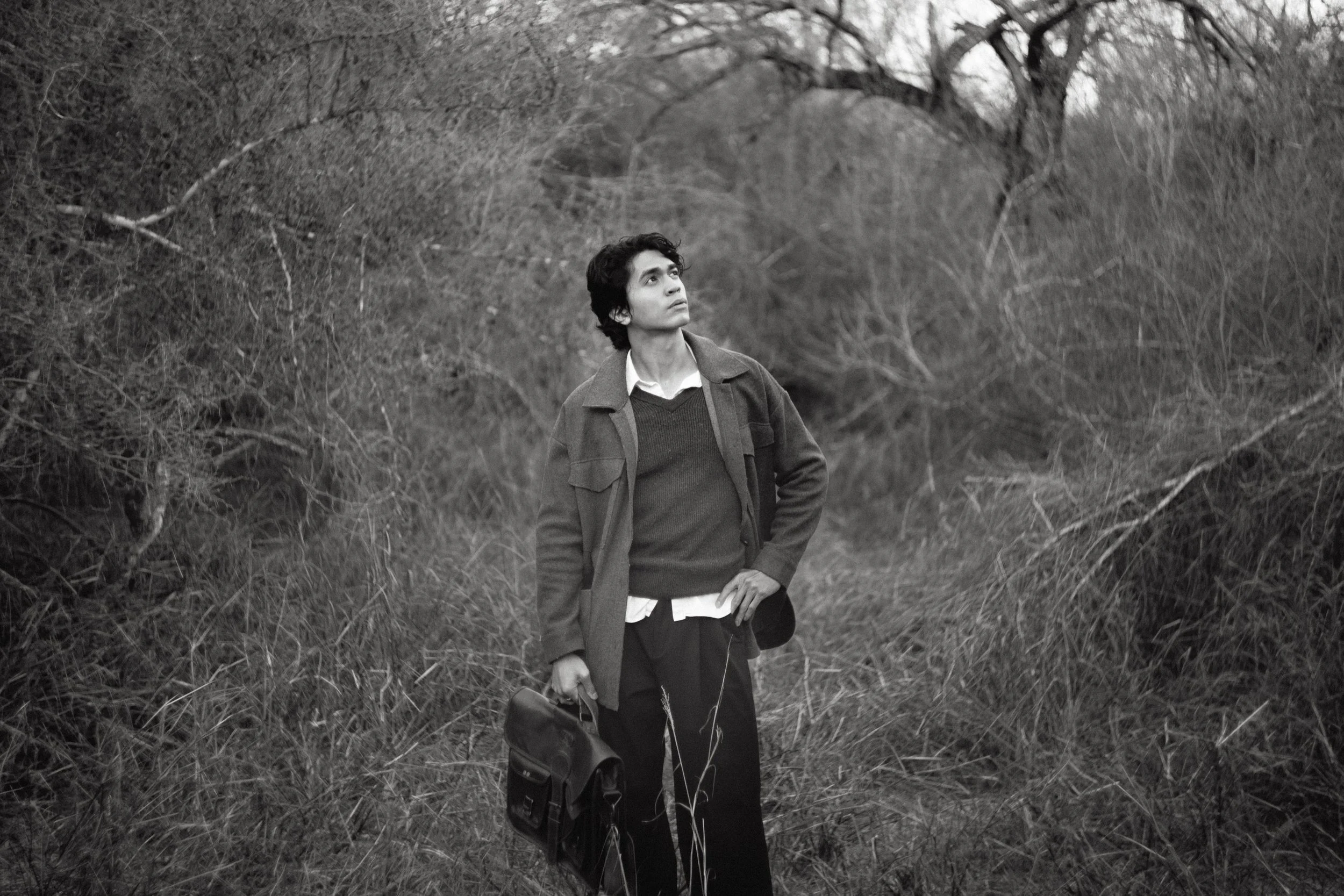 A young man standing in a wooded area, looking upward, carrying a briefcase in his right hand and a bag over his shoulder, dressed in casual work attire.