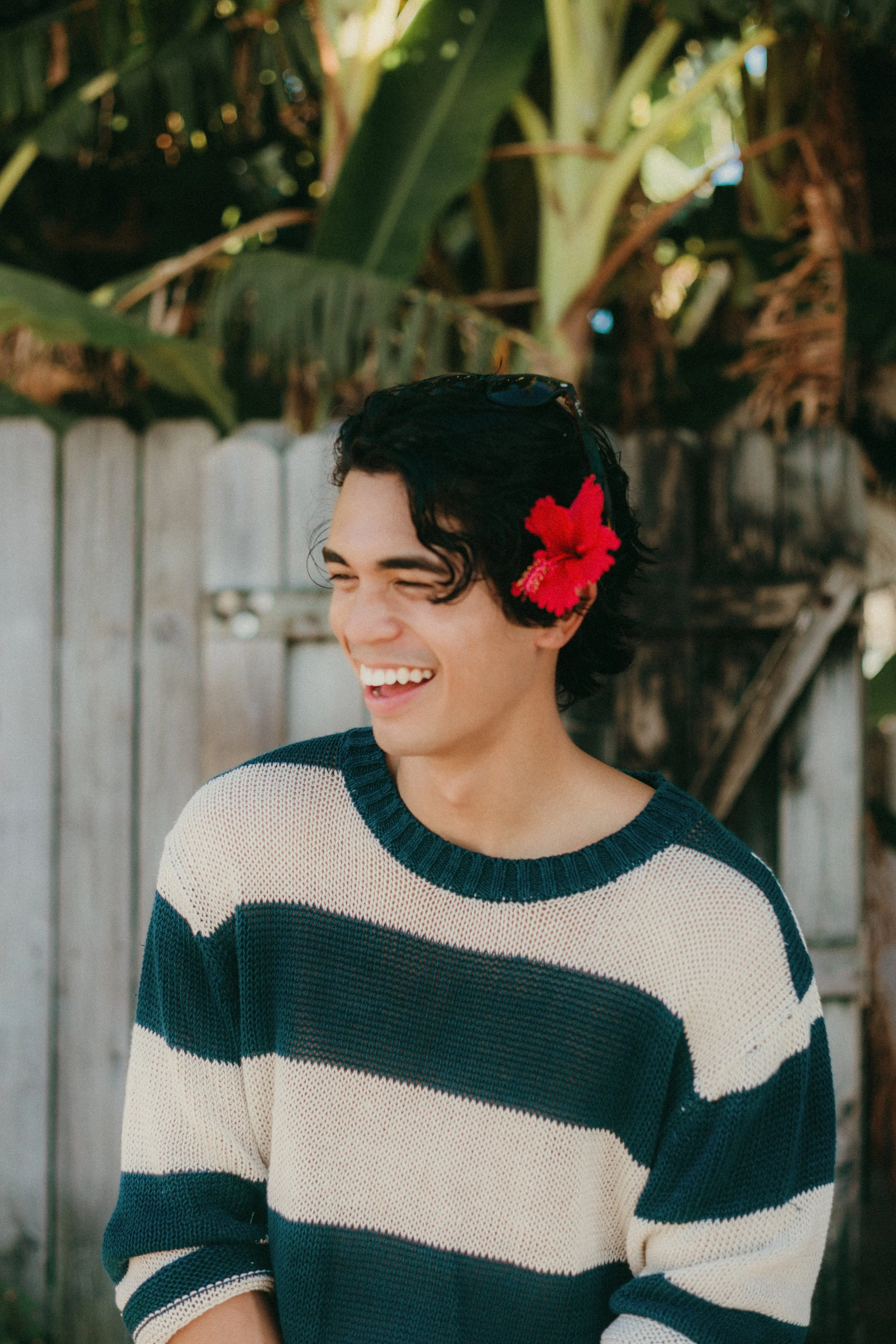 A young man with dark curly hair wearing a striped sweater, smiling and looking away, with a red hibiscus flower behind his left ear, standing outdoors in front of a wooden fence and tropical plants.
