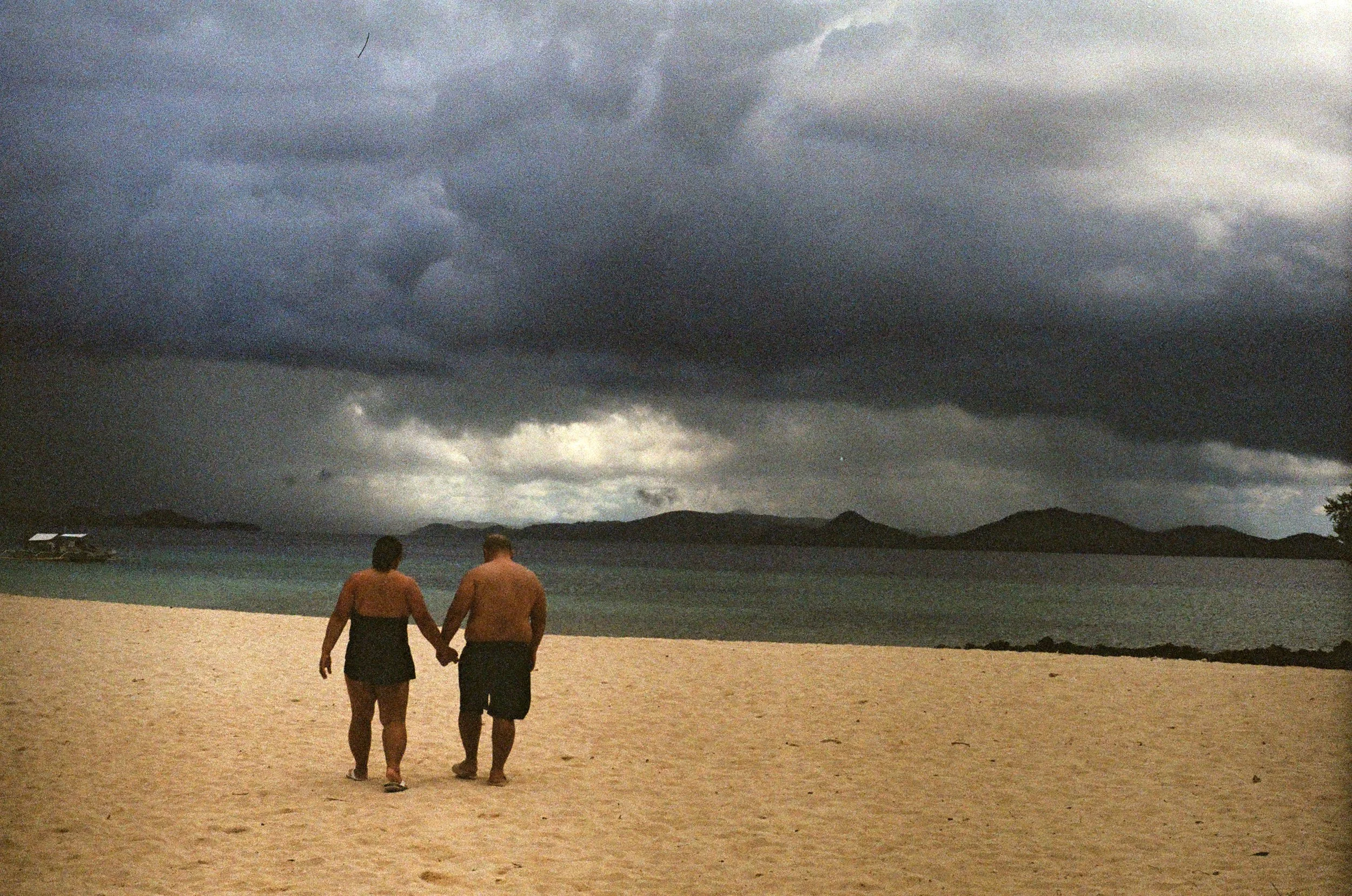 A couple walking on a sandy beach holding hands under a cloudy, stormy sky with mountains in the distance.