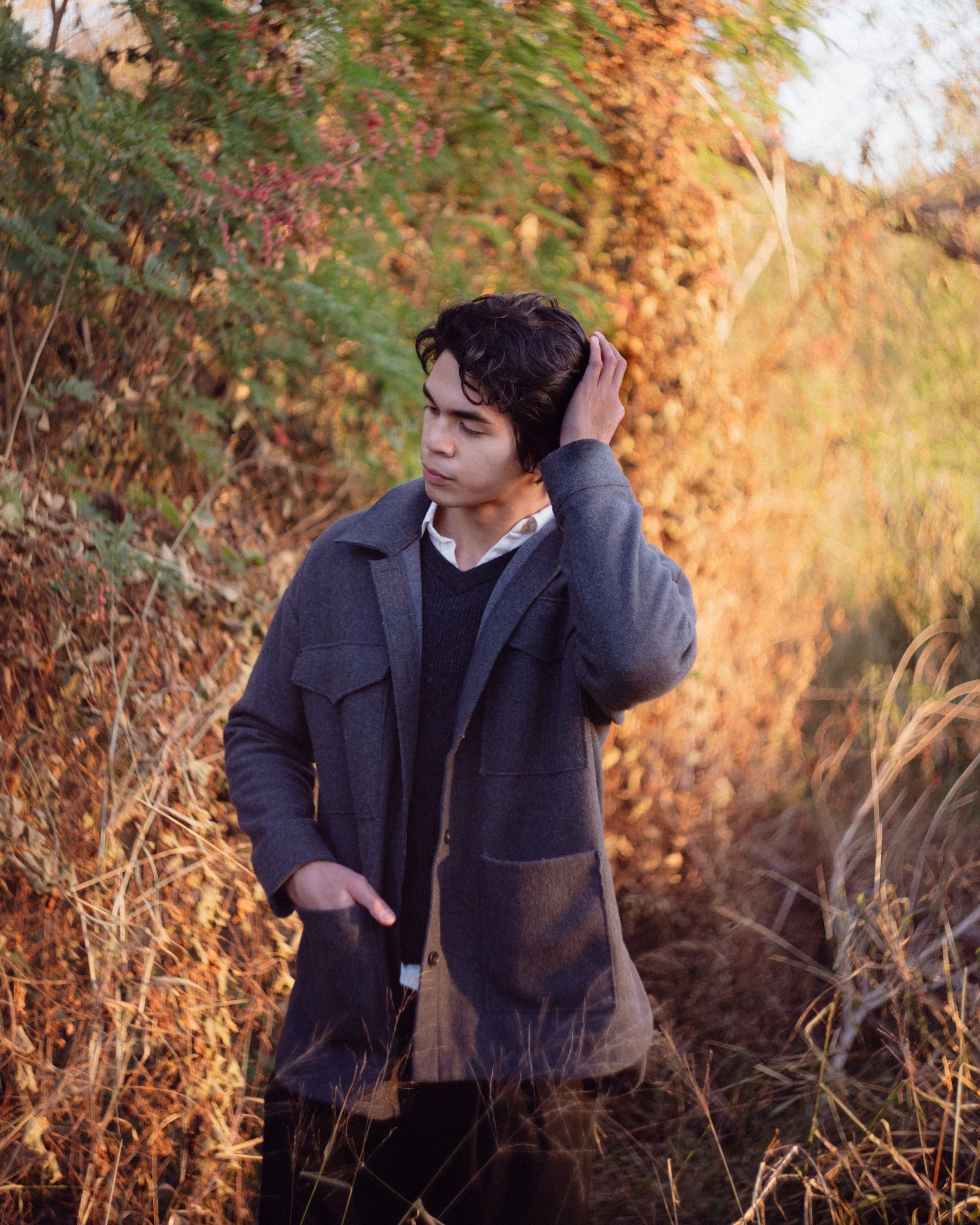 A young man in a dark gray coat standing outdoors among autumn foliage, resting his hand on the back of his head and looking down thoughtfully.