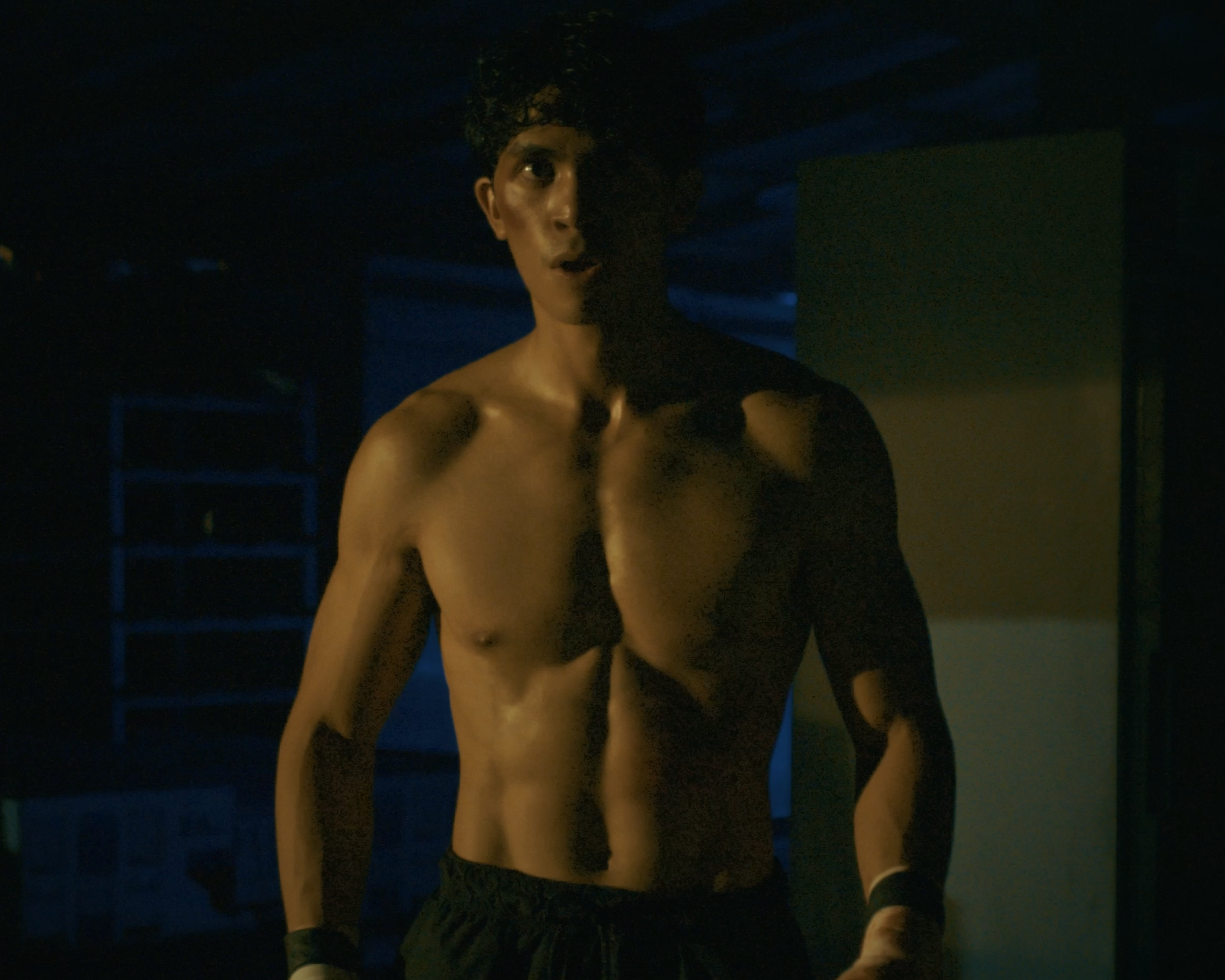 A shirtless young man with curly hair standing in a dimly lit gym, with a surprised expression on his face.