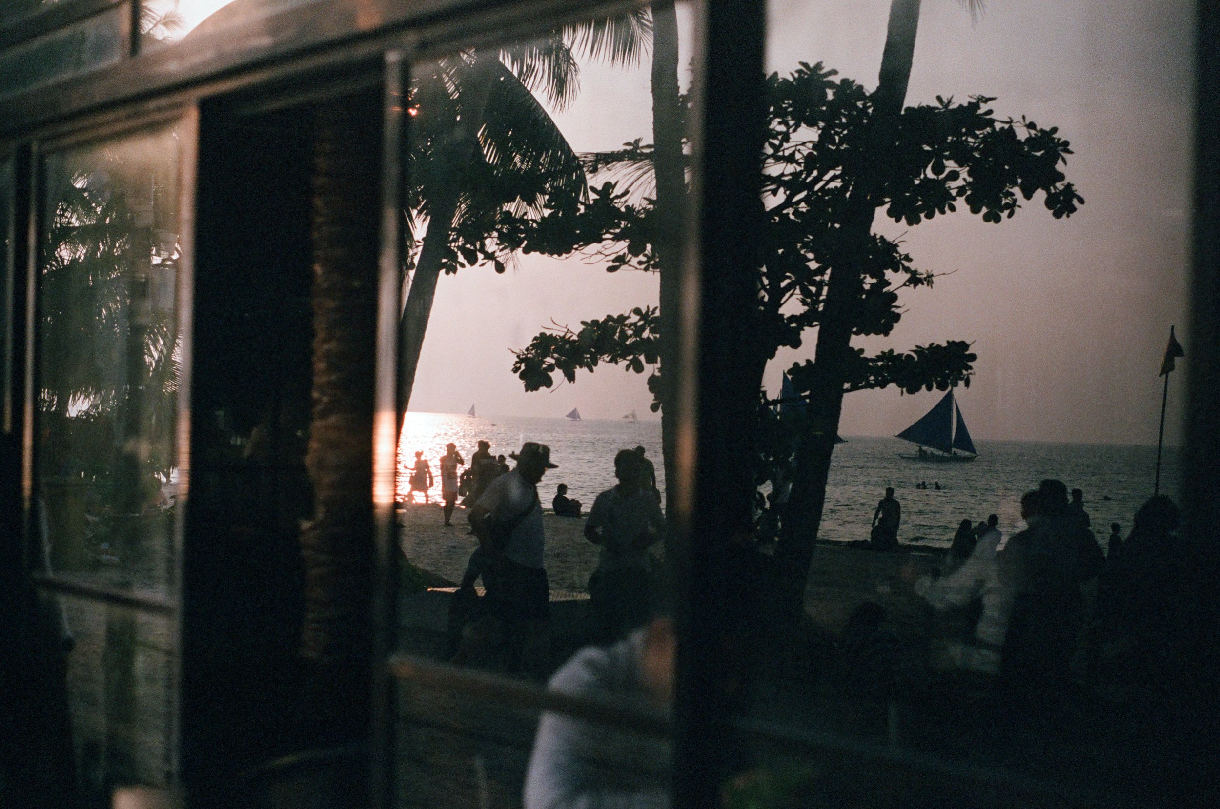 People gathered on a beach at sunset, silhouetted by the evening sky, with sailboats on the water and trees framing the scene.