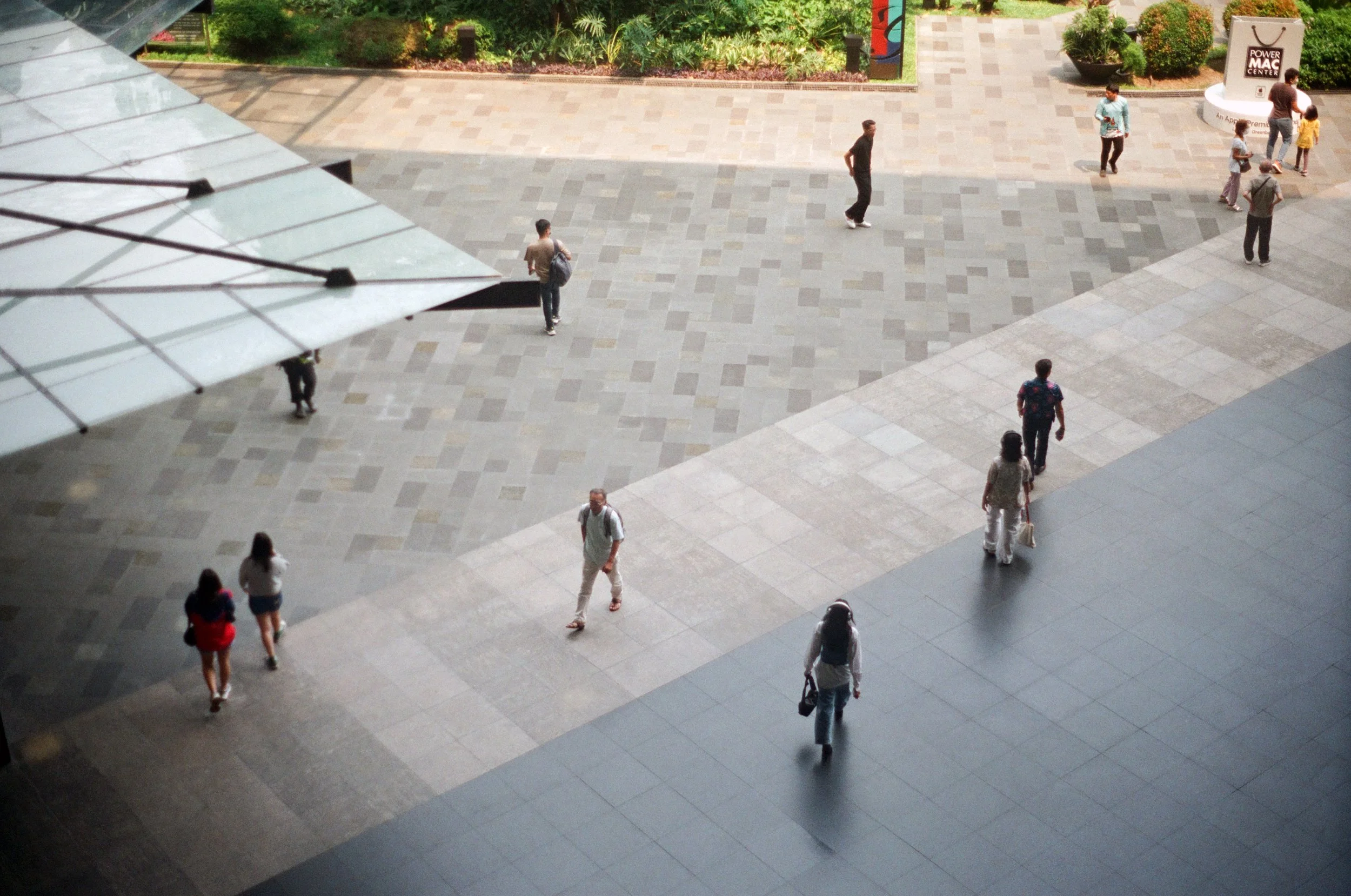 People walking in an outdoor shopping or public area, with trees and planters, and a large sign reading 'Power Mac Center' in the background.