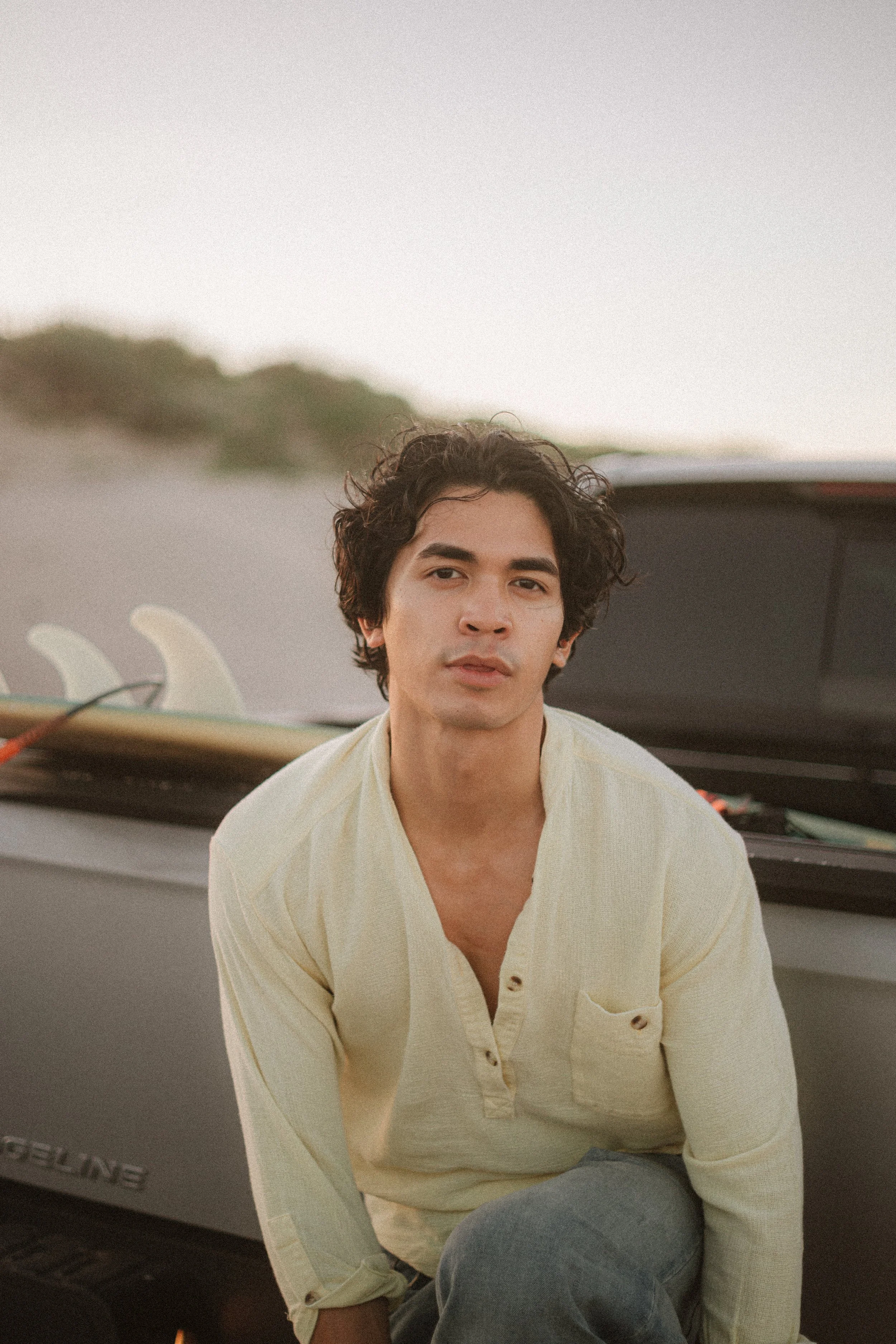 A young man with dark, curly hair and light skin, wearing a light yellow shirt and blue jeans, sitting outdoors near a boat with surfboards in the background.