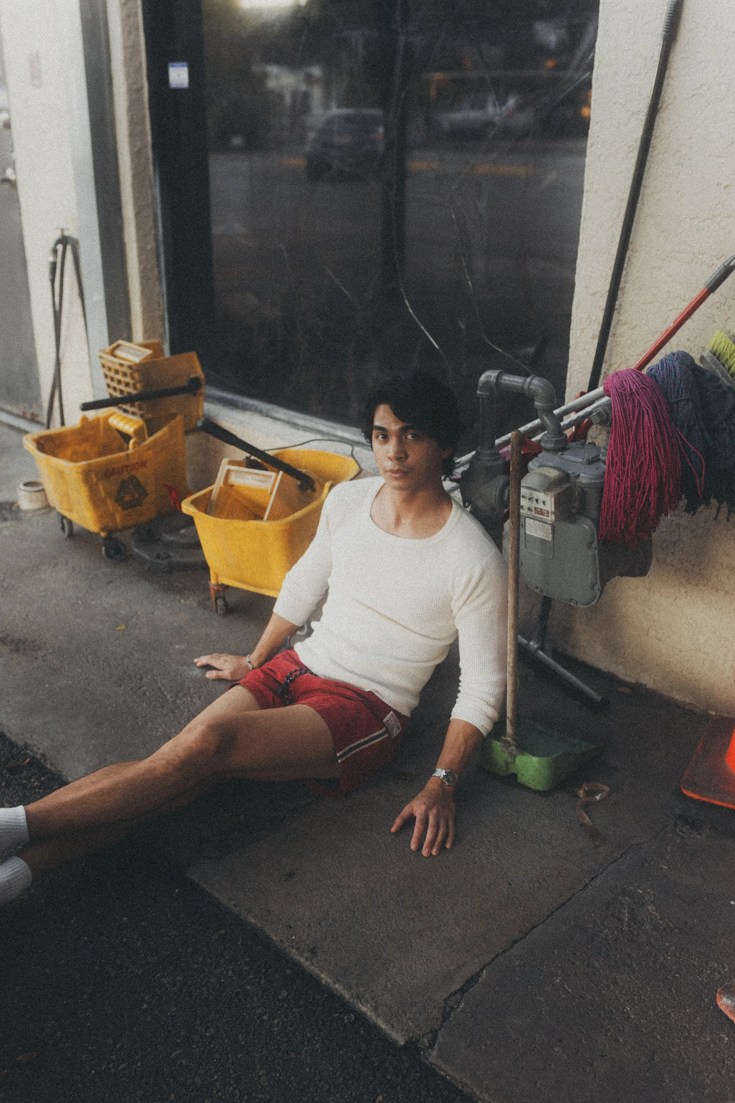 A young man with black hair, wearing a white long-sleeve shirt and red shorts, sitting on the sidewalk next to cleaning supplies, including yellow mop buckets, mops, and a cleaning cart, outside a building with a large window.