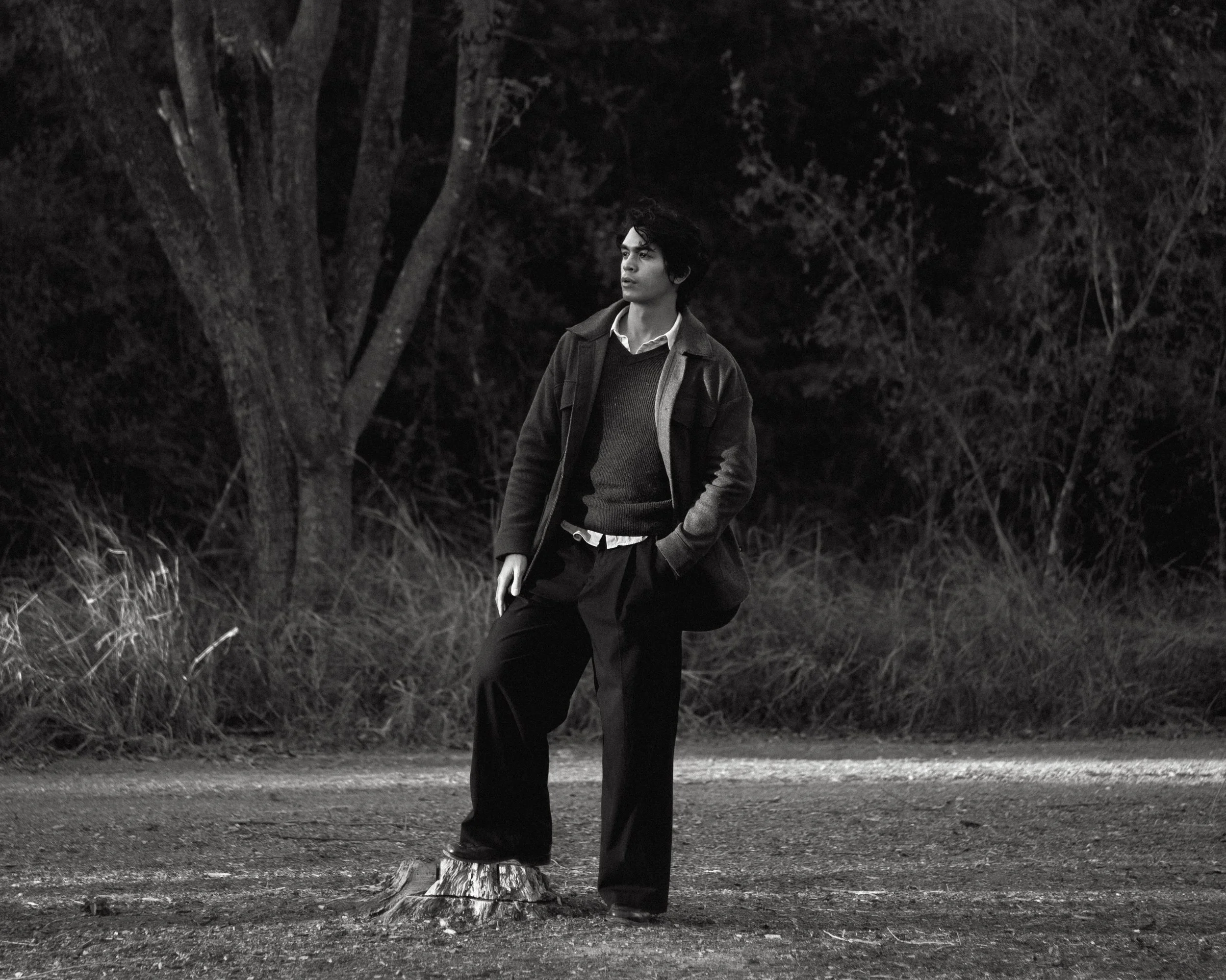 A young man with dark curly hair dressed in a layered outfit including a sweater, shirt, and jacket, standing outdoors with one foot on a tree stump, against a backdrop of trees and foliage in black and white.