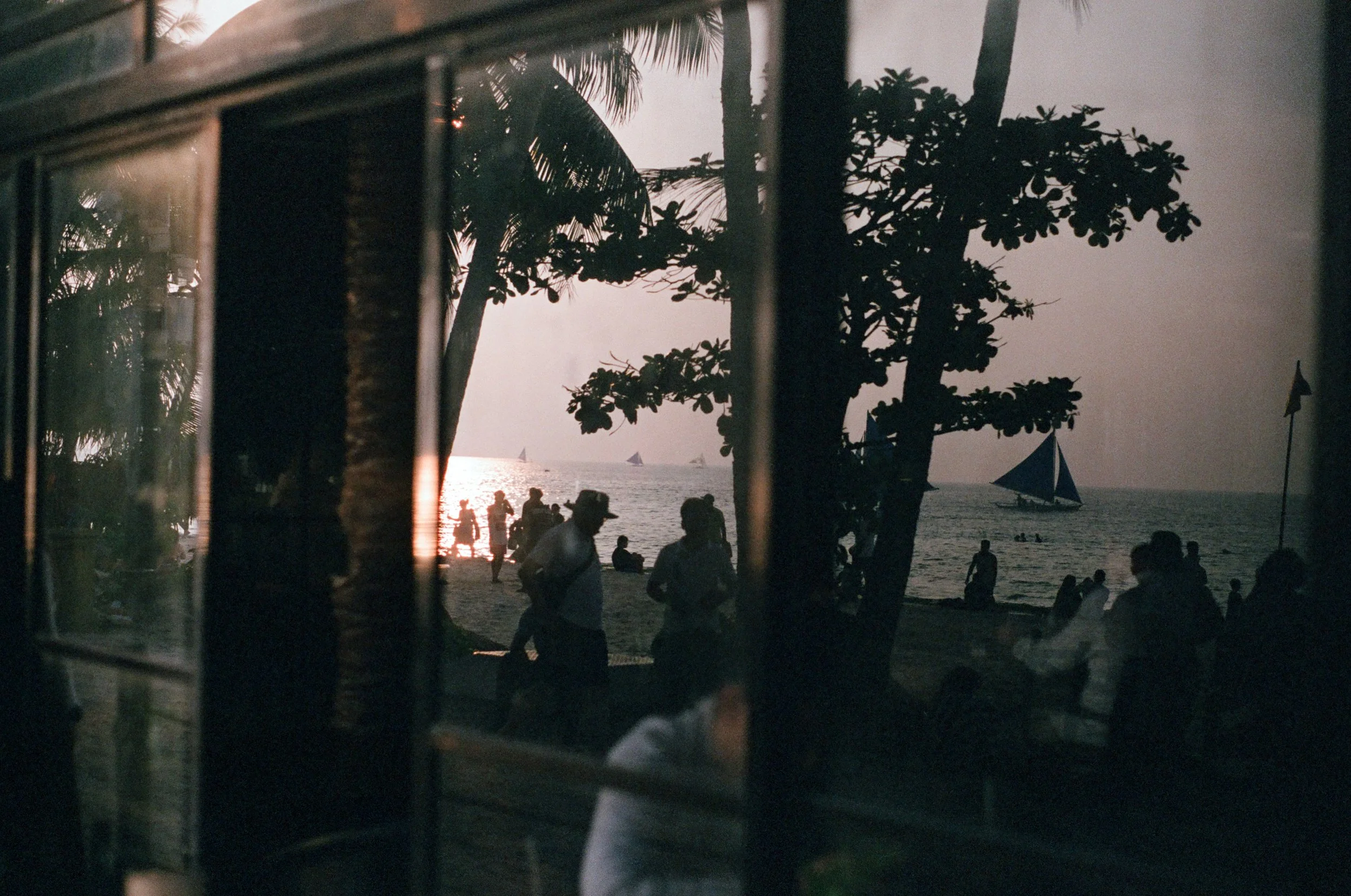 People on a beach viewed through a window during sunset with sailboats on the water and trees silhouette.