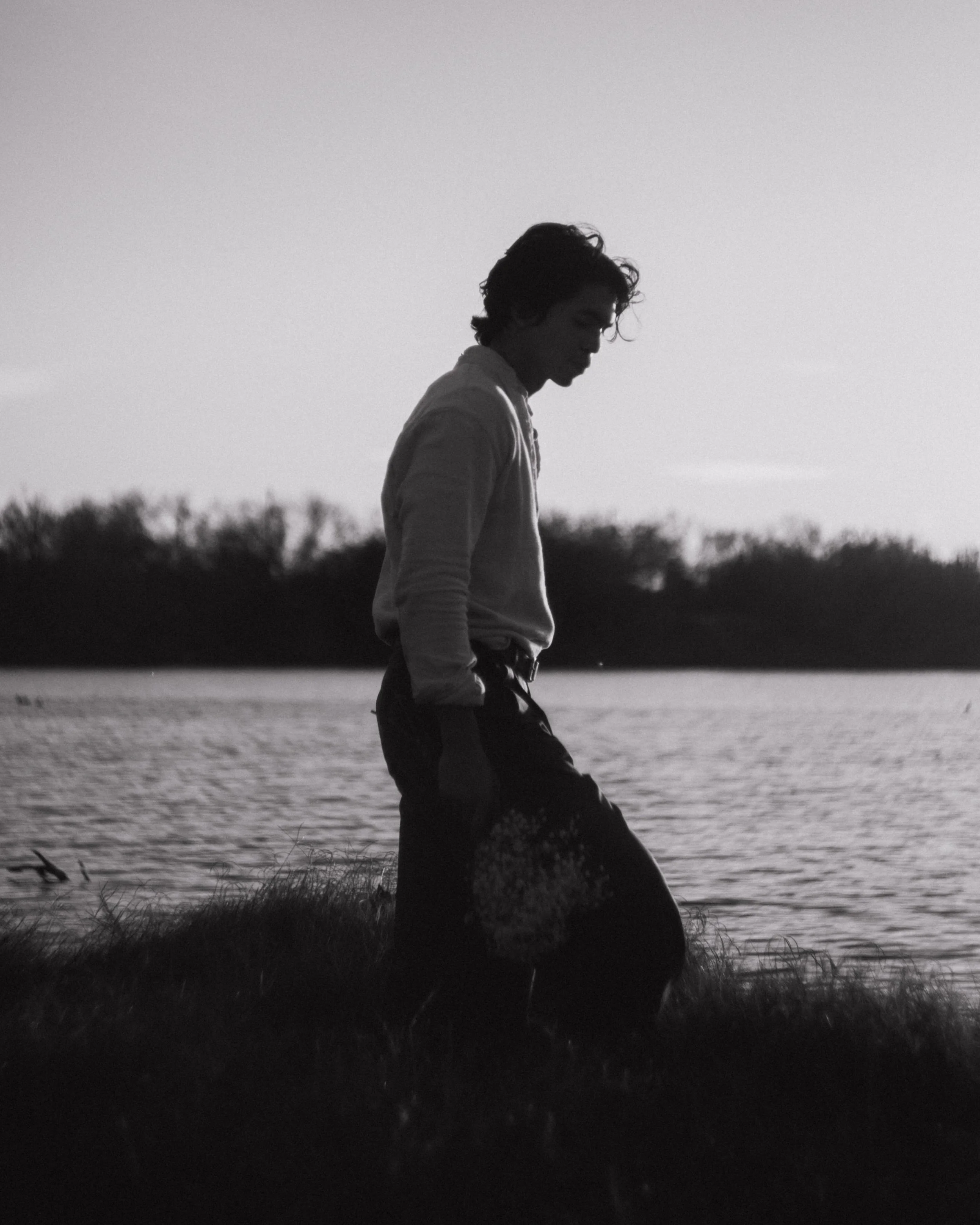 A person standing on grass near a body of water during sunset, in black and white.