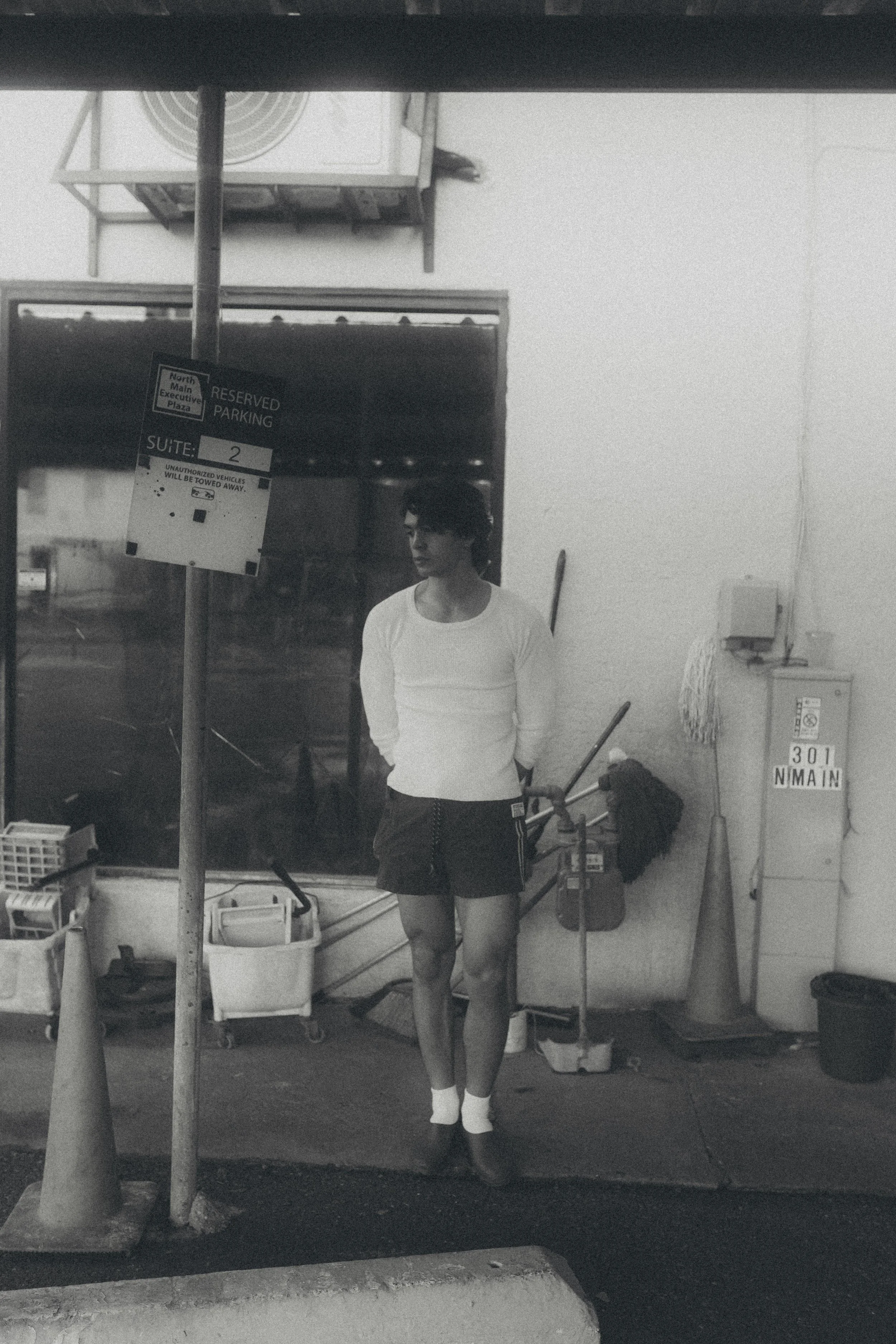 Black and white photo of a woman standing in a doorway of a commercial building, with cleaning supplies, a mop, and cleaning cart nearby.