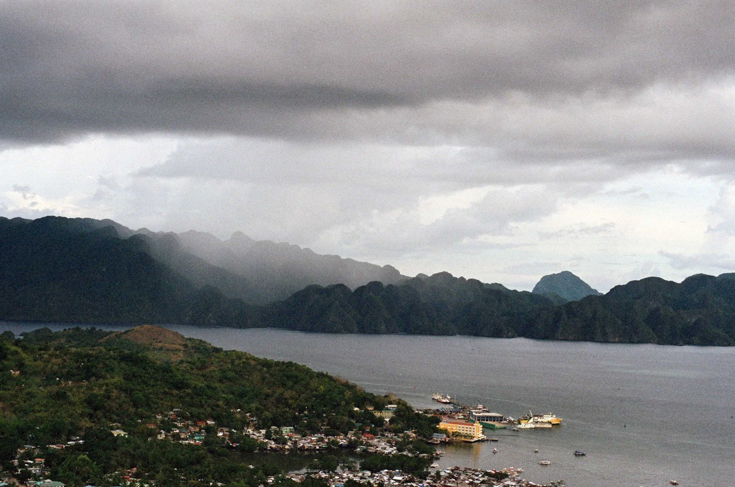 A coastal town along a bay with boats docked at the pier, surrounded by green hills and mountains under a cloudy sky.