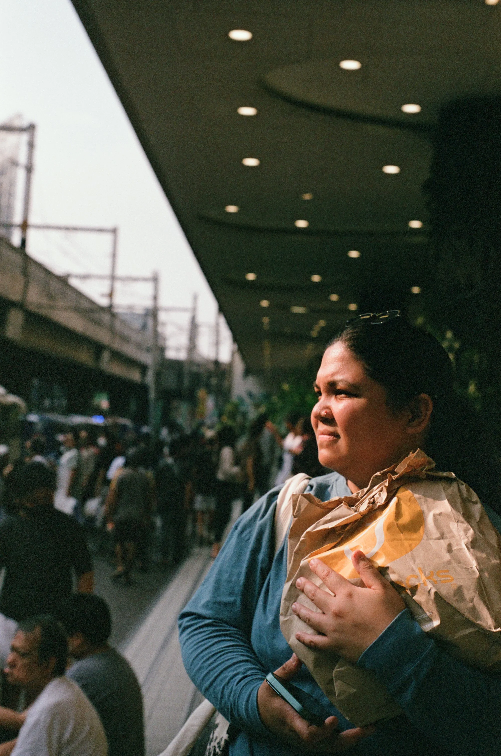 A woman holding a brown paper bag and a smartphone at an outdoor public area, with a crowd of people and an overpass in the background.