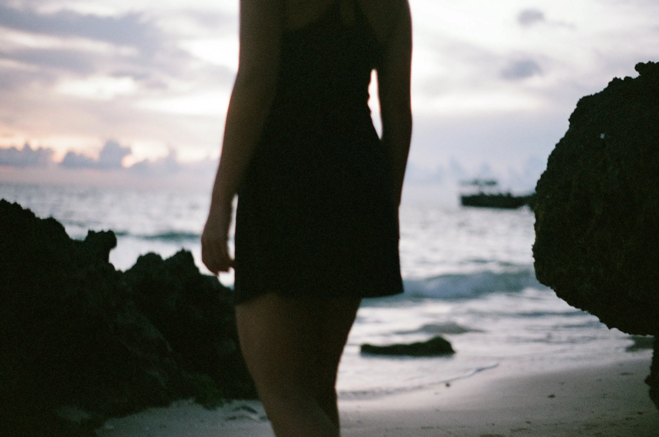 A person in a black dress standing on a beach between rocks during sunset, with the ocean and a boat in the background.