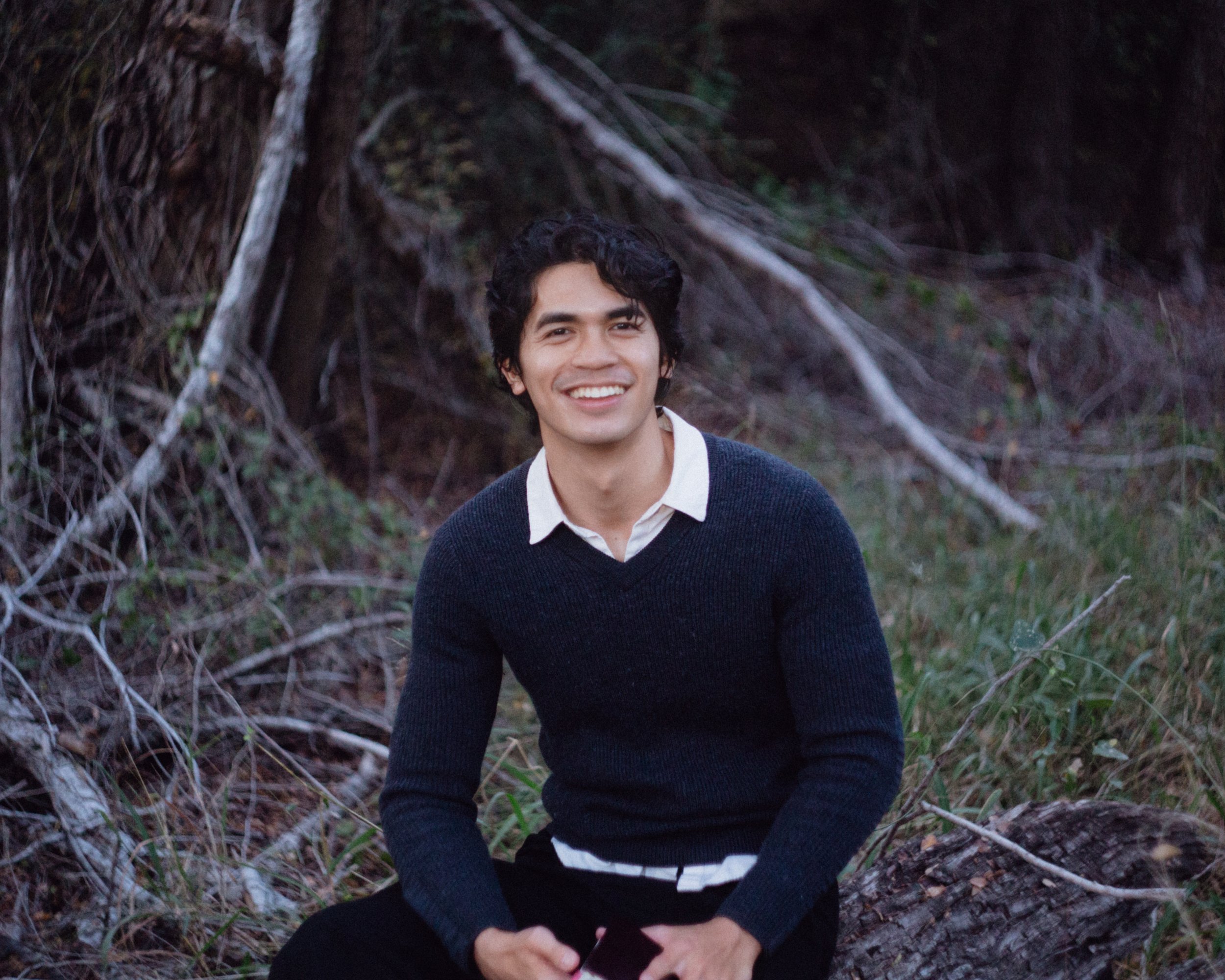 A young man with curly dark hair, wearing a white shirt and dark sweater, smiling and sitting outdoors on a fallen log in a wooded area.