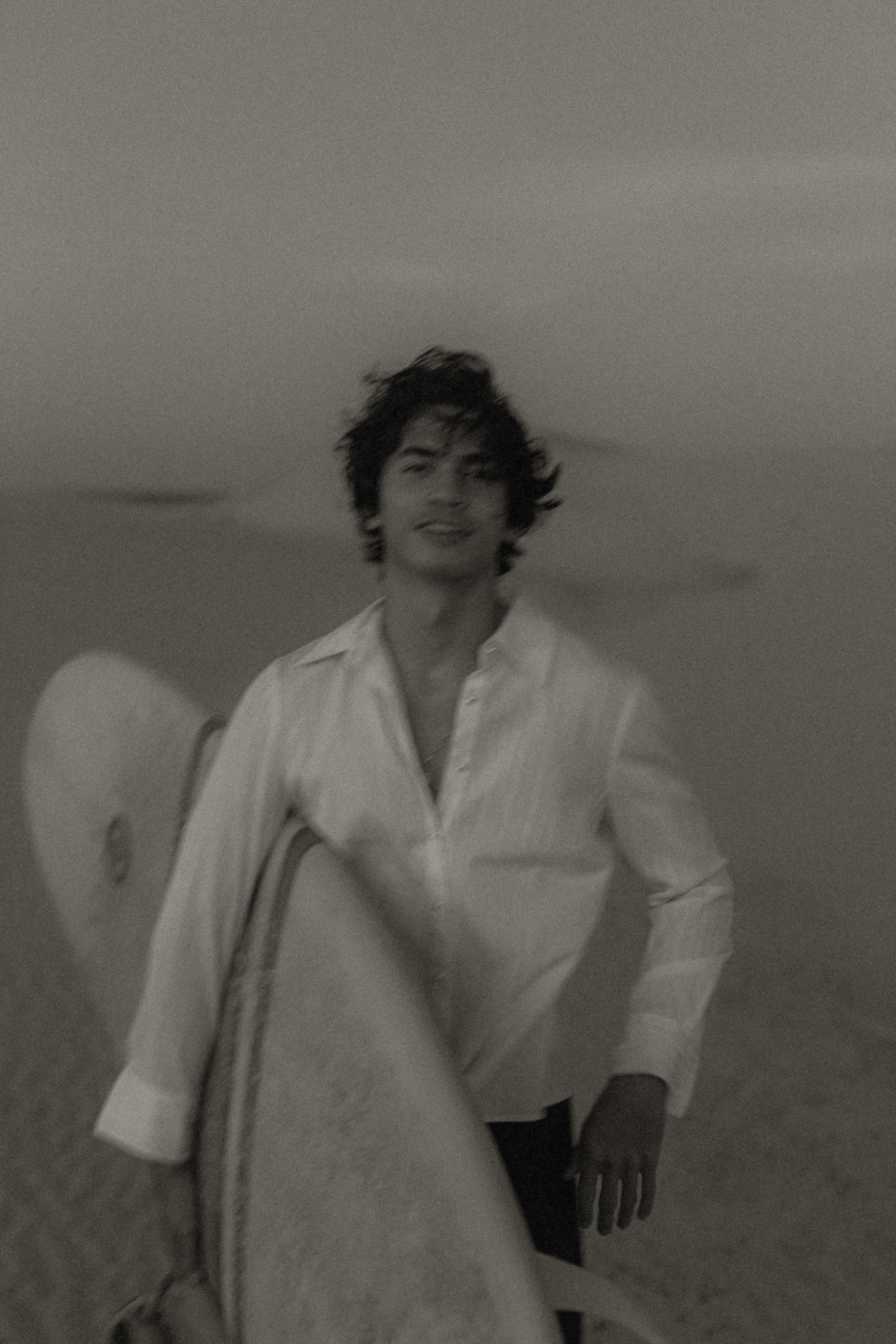 A woman with curly hair wearing a white button-up shirt, holding a surfboard on a beach.