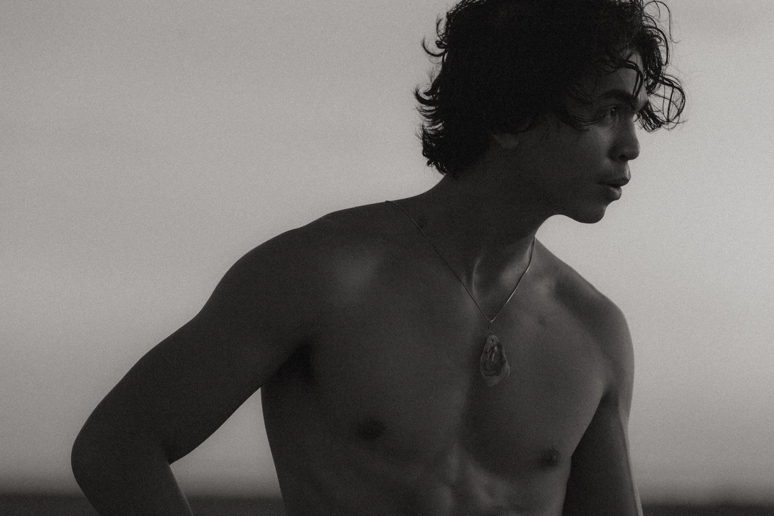 A black-and-white photo of a shirtless young man with curly hair, looking to the side, wearing a pendant necklace.