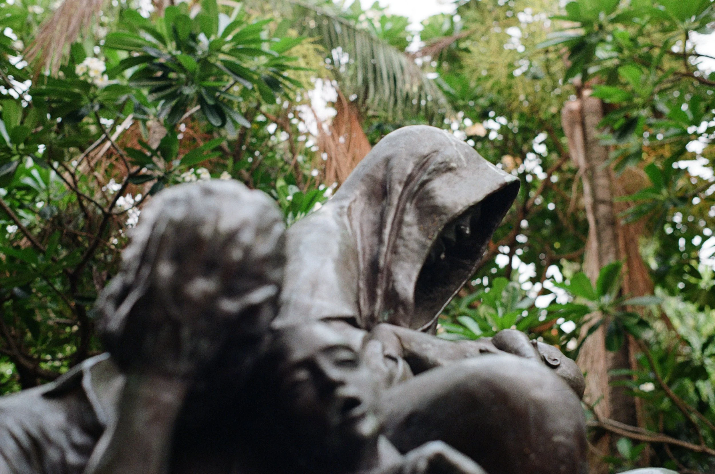 Close-up of a statue of a woman with her mouth open, holding a child, surrounded by lush green foliage and trees in the background.