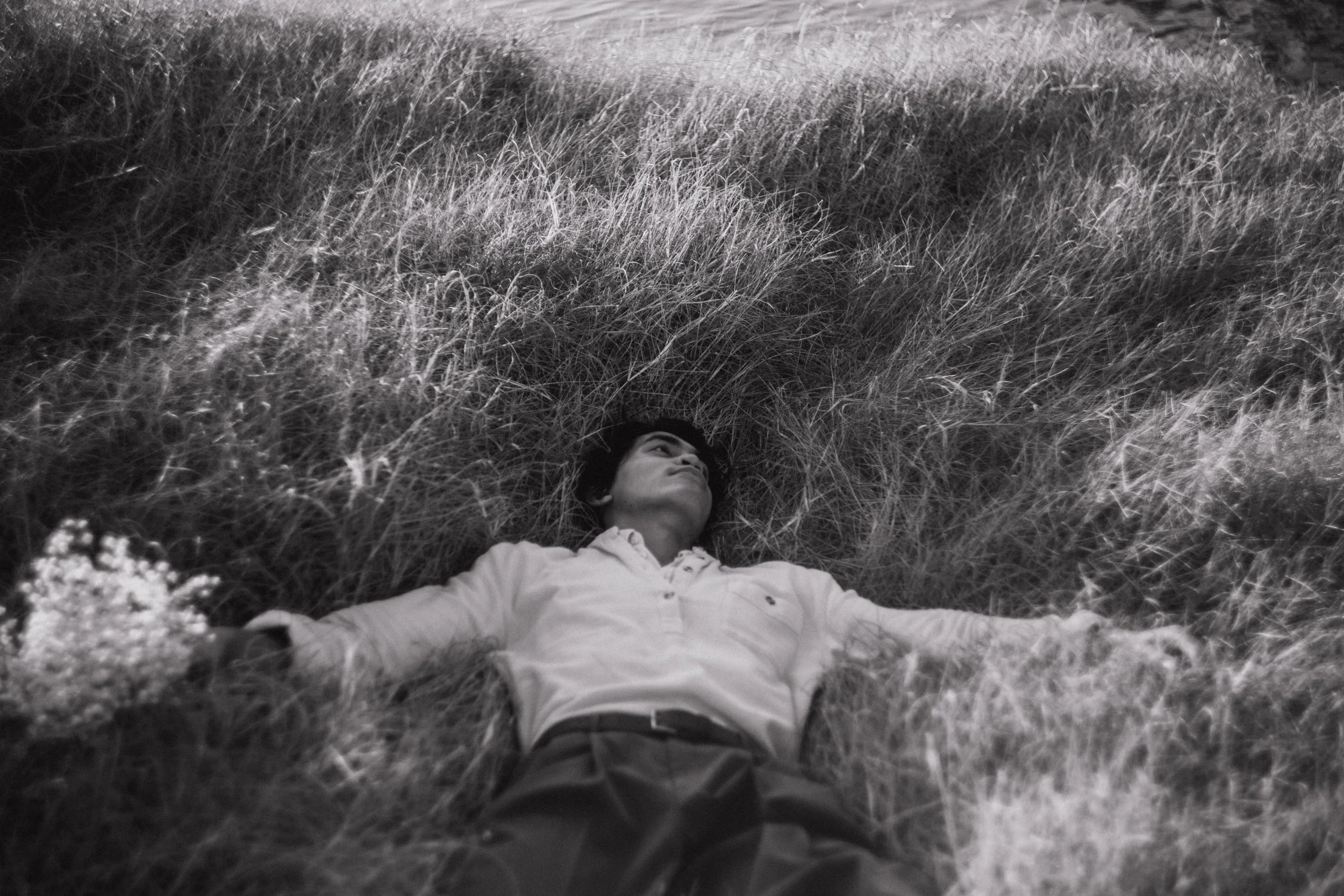 A man lying on his back in tall grass, with arms outstretched and looking upward.