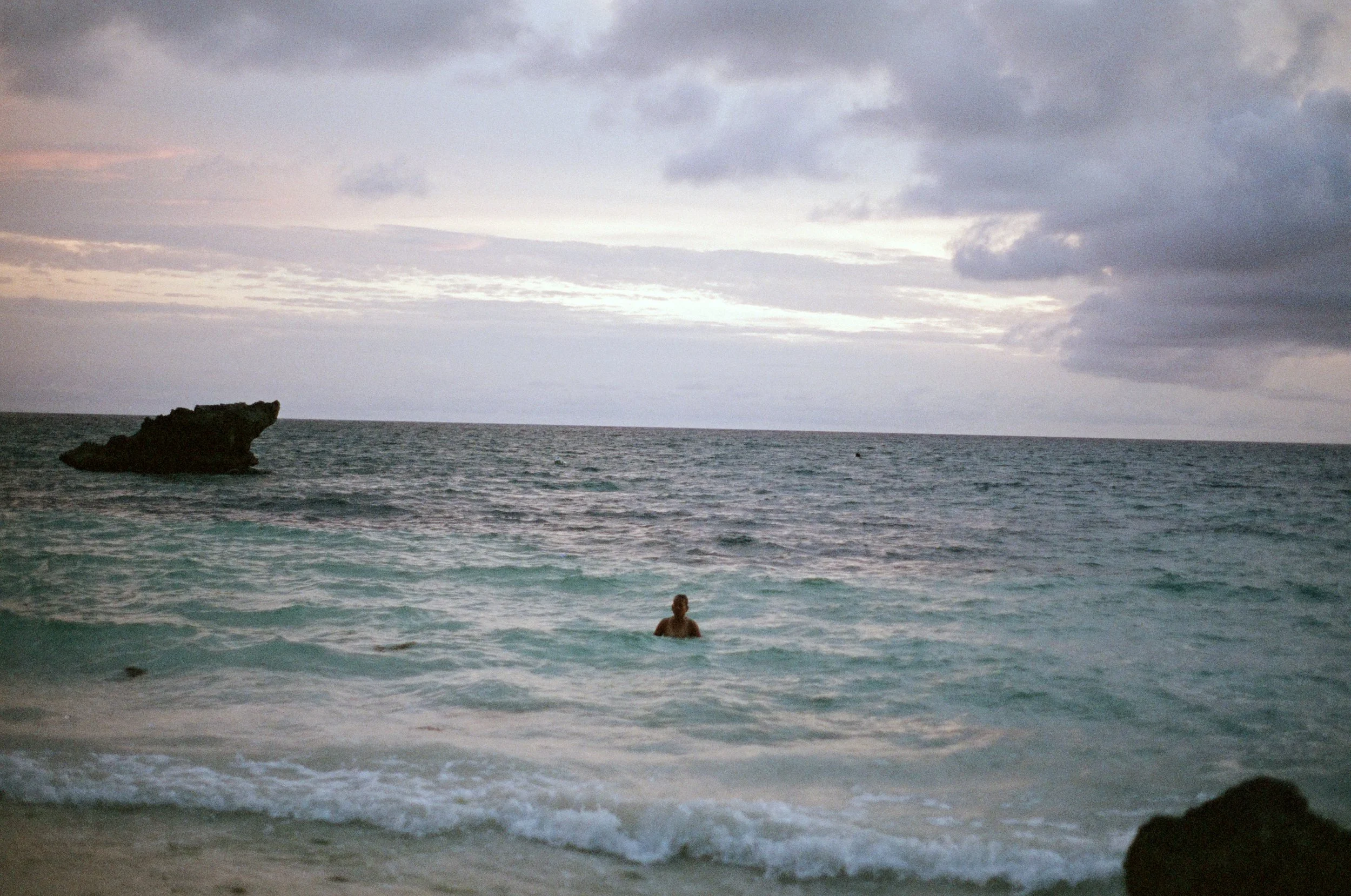 A person swimming in the ocean near a sandy beach with a large rock formation in the distance under a cloudy sky during sunset or sunrise.