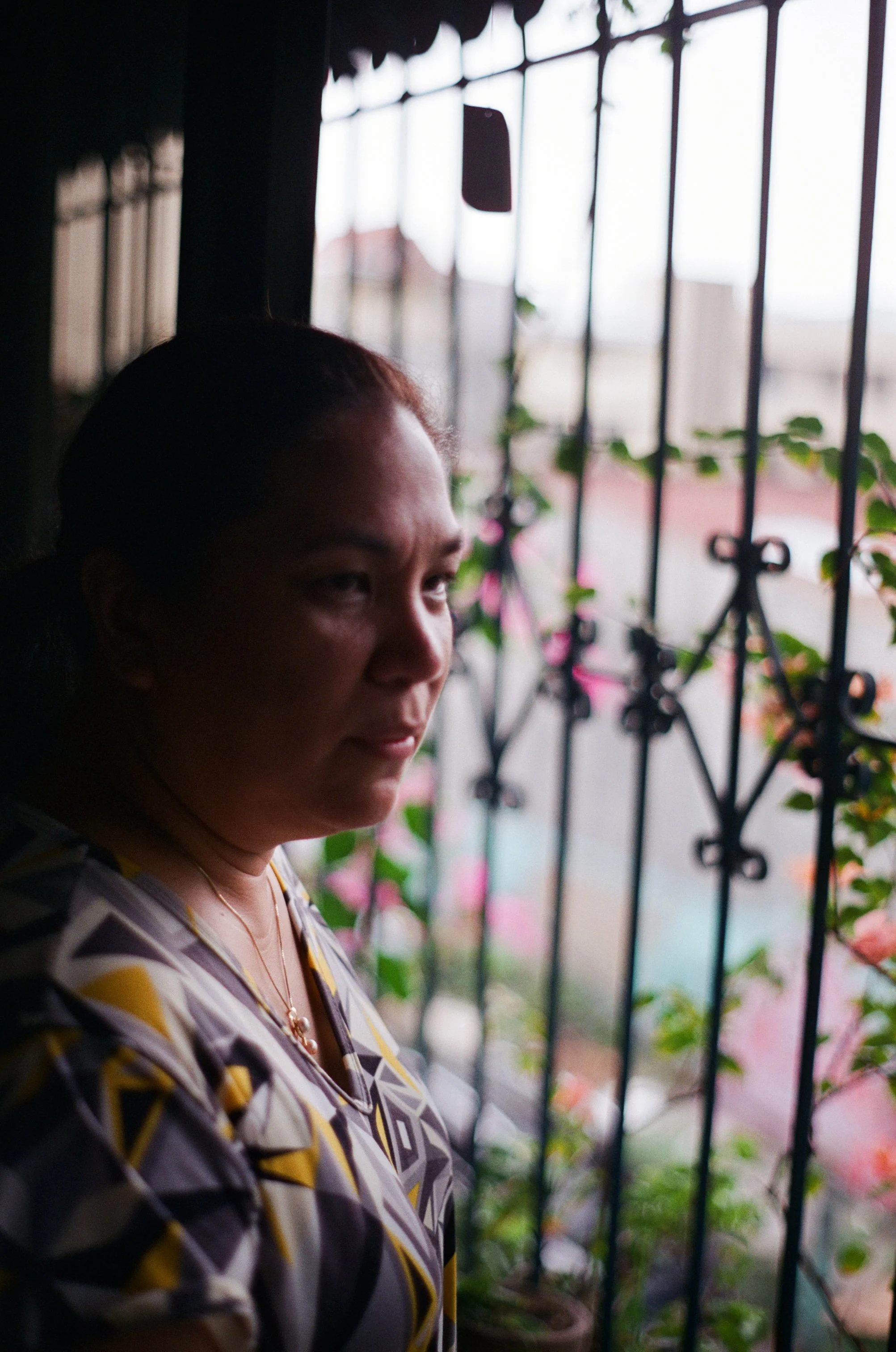 A woman stands near a window with black iron bars and pink flowers outside. She has dark hair pulled back and wears a patterned shirt with yellow, black, and white colors.
