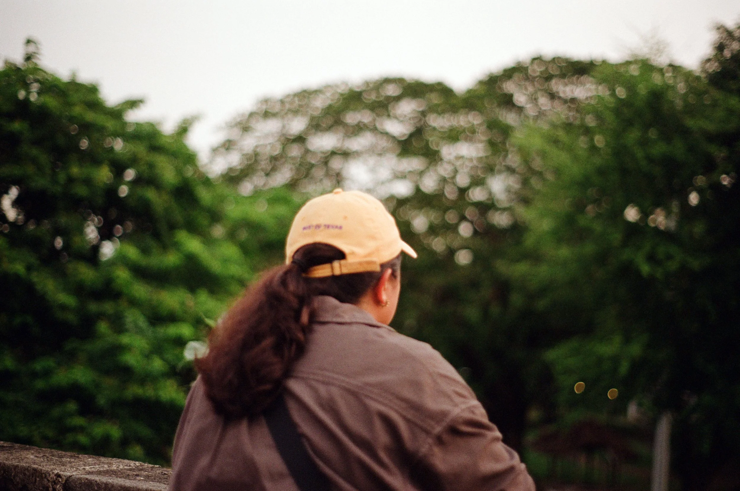 A woman wearing a peach-colored cap and brown jacket sitting outdoors with her back to the camera, surrounded by green trees.