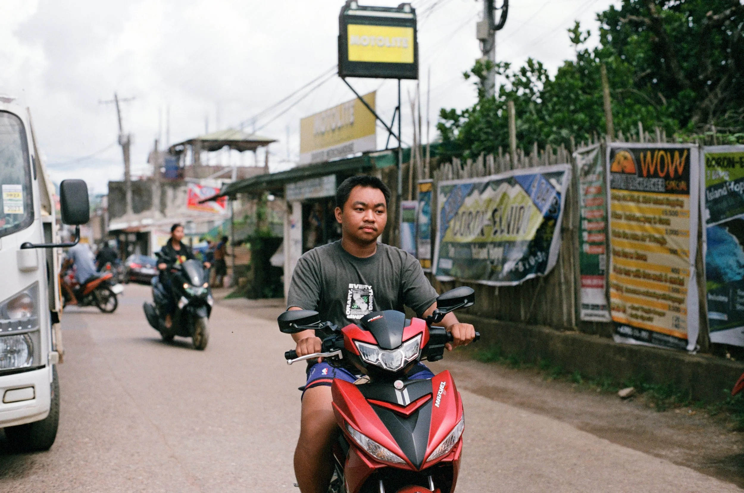 A young man riding a red motorcycle on a busy street with other motorcyclists and vehicles passing by. There are shops and signs along the roadside.