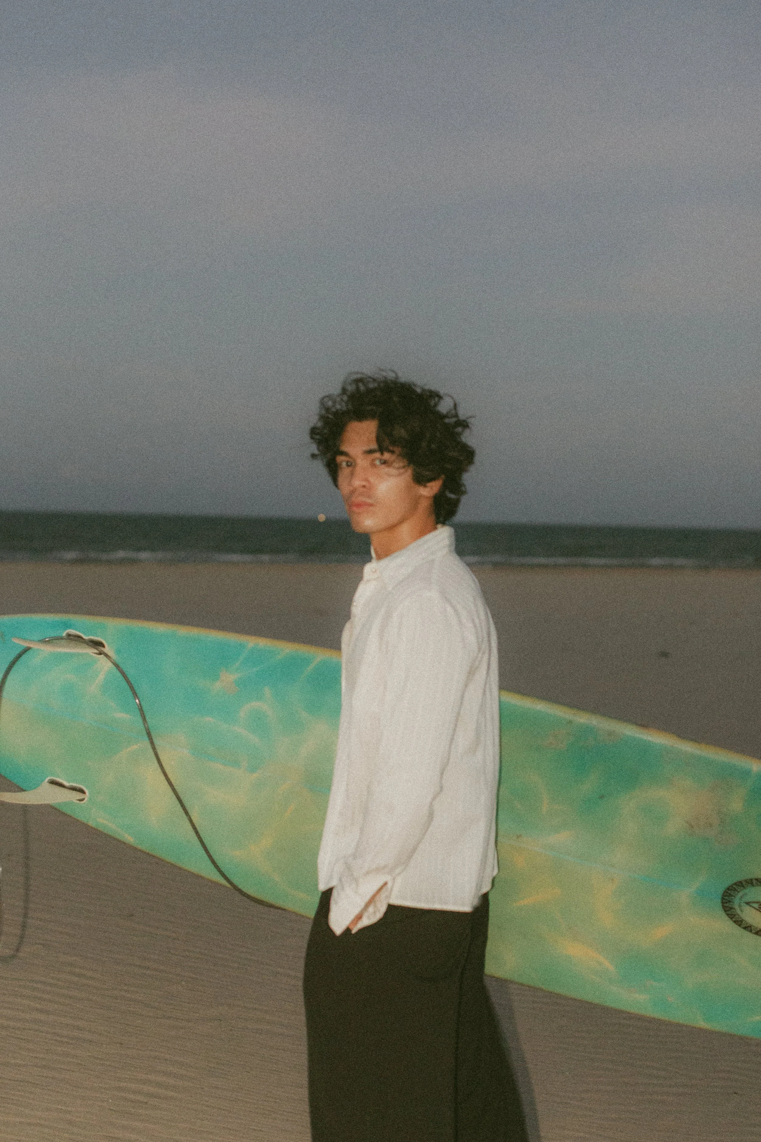 A man with curly dark hair, wearing a white shirt and dark pants, stands on a beach at dusk holding a surfboard with a psychedelic design.