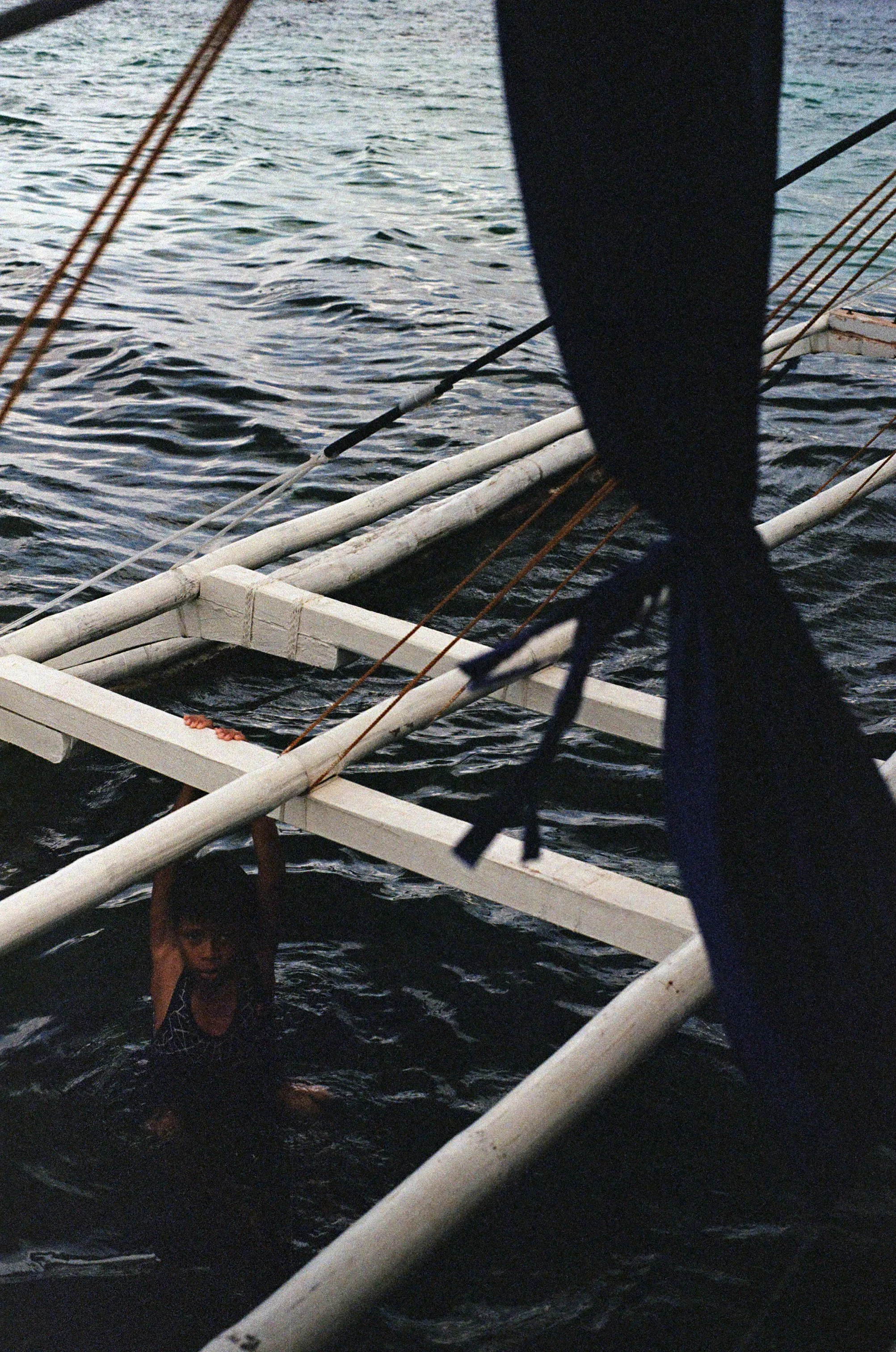 A young girl in a swimsuit swimming near a white boat frame and black sail on a body of water.