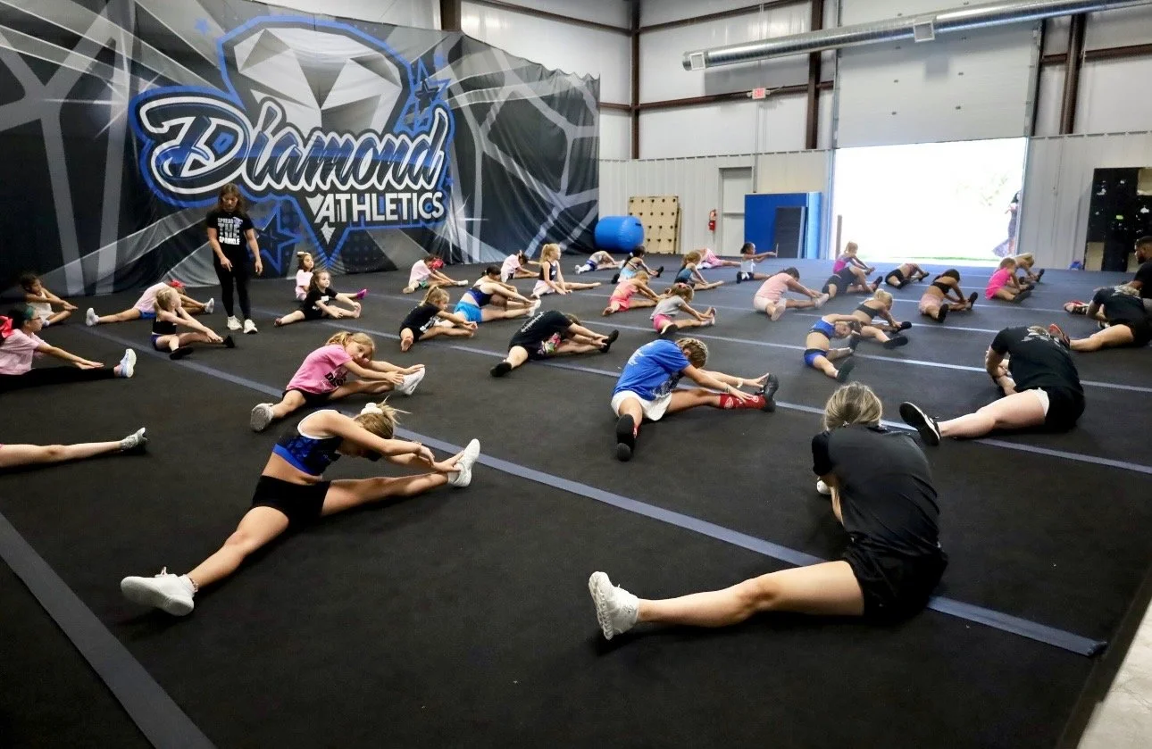 Young children in a gymnastics class stretching on a black mat inside a gym with a large wall mural that reads 'Diamond Athletics' and features a diamond graphic.