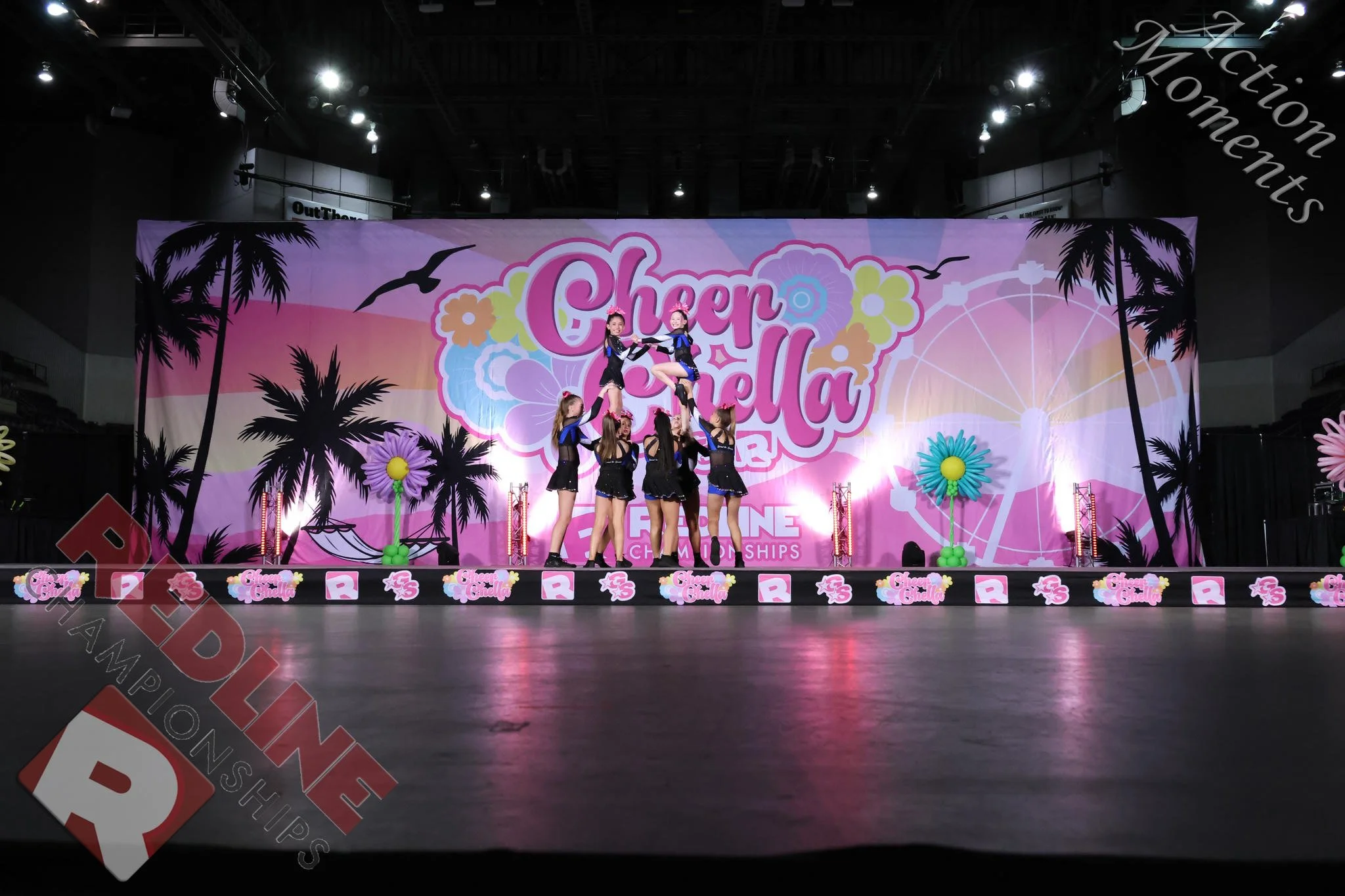 Cheerleaders performing a stunt on stage at a cheer and dance championships with a colorful backdrop featuring palm trees and flowers.