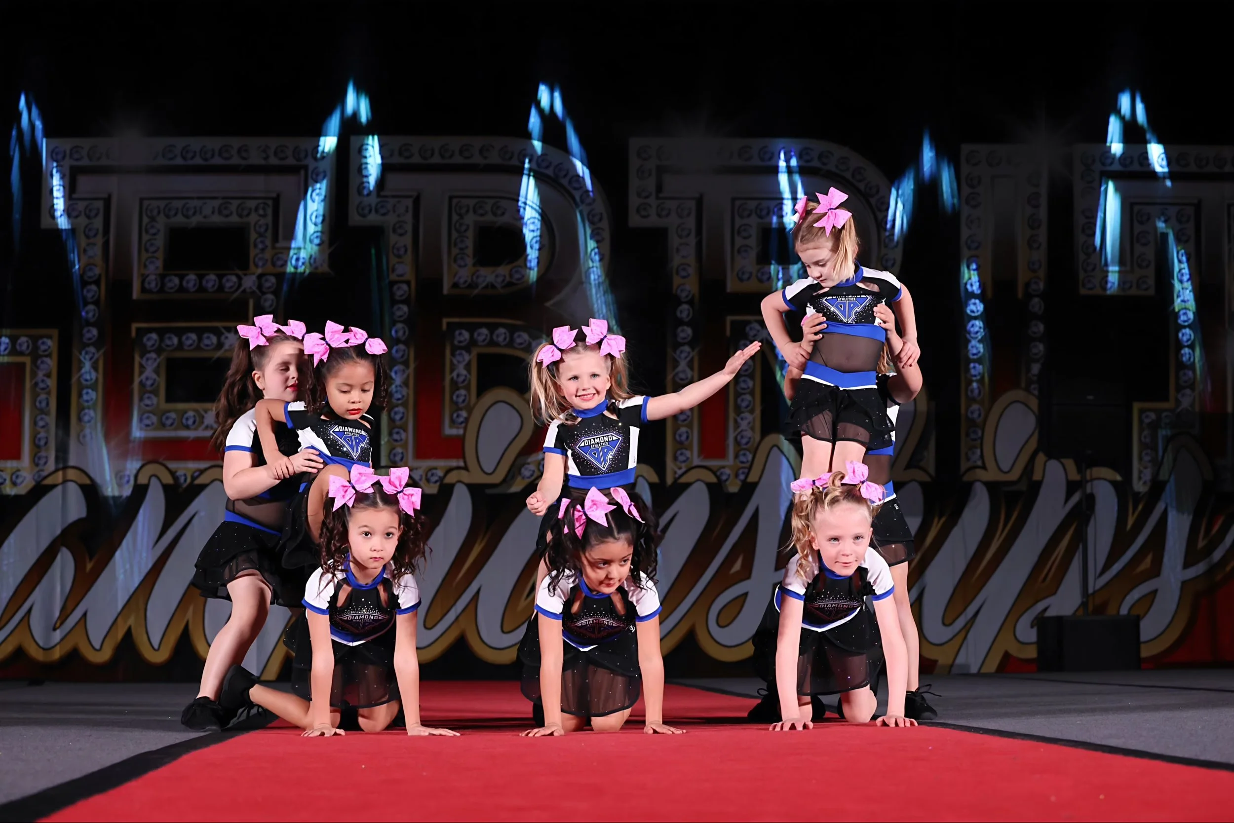 Group of young girls performing a cheerleading or dance routine on stage, some kneeling and some standing, wearing black and blue costumes with pink bows in their hair.