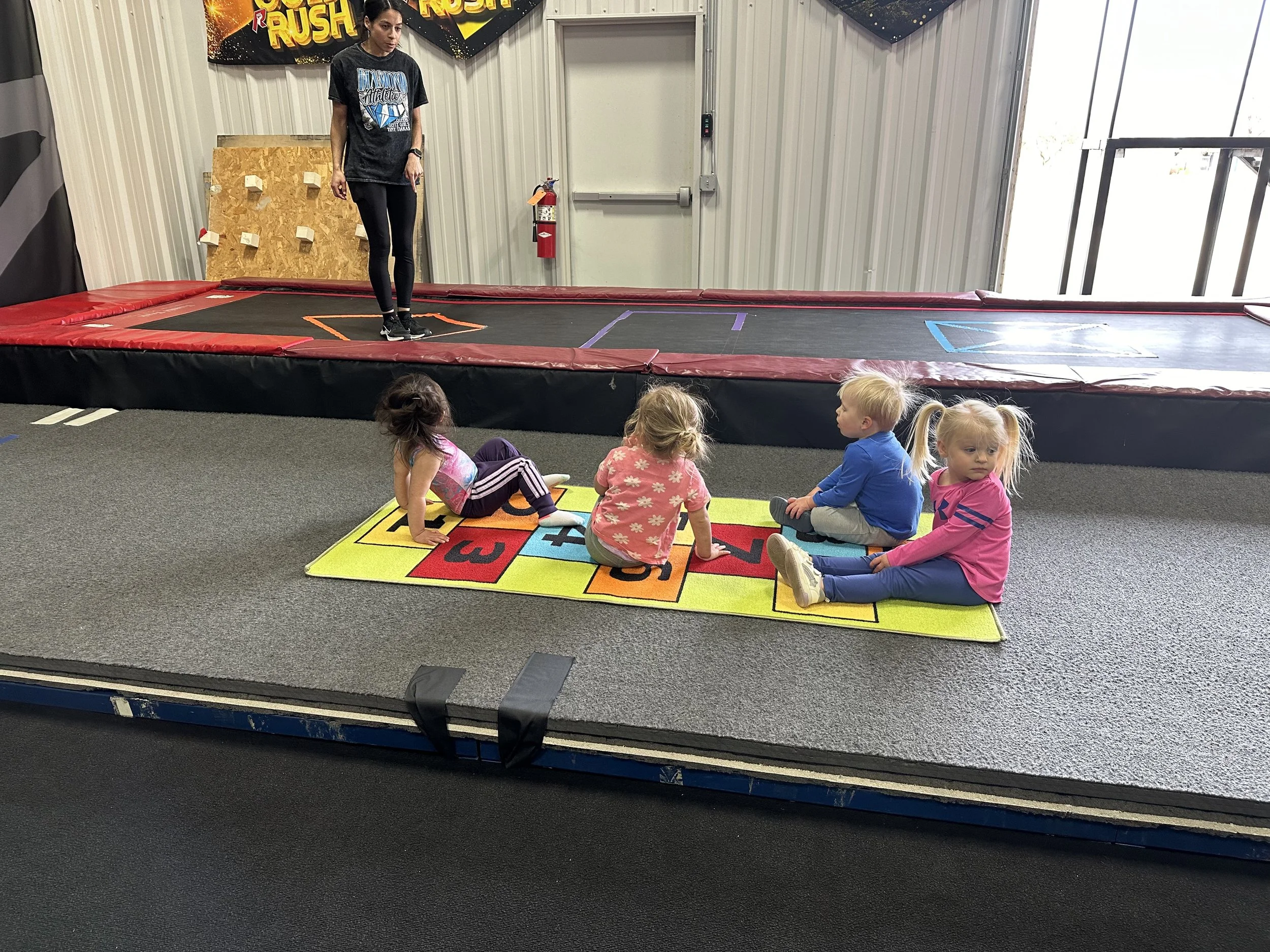 A group of children sitting on a colorful number rug while a woman gives instructions on a trampoline at an indoor play area.