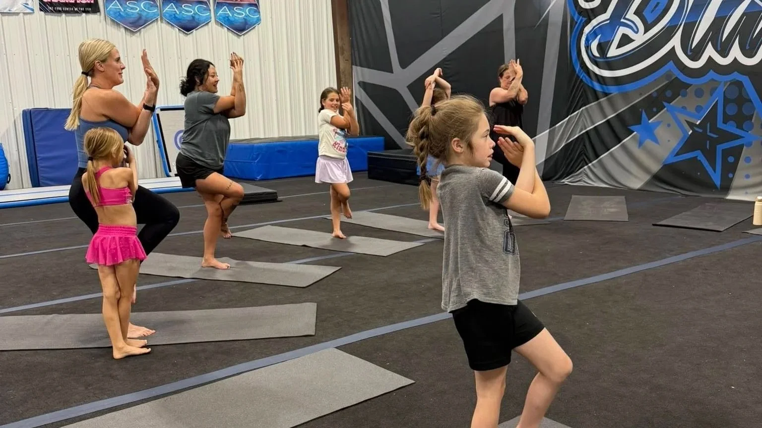 A group of children and an adult participating in a gymnastics class in a gym, practicing stretching poses on mats.